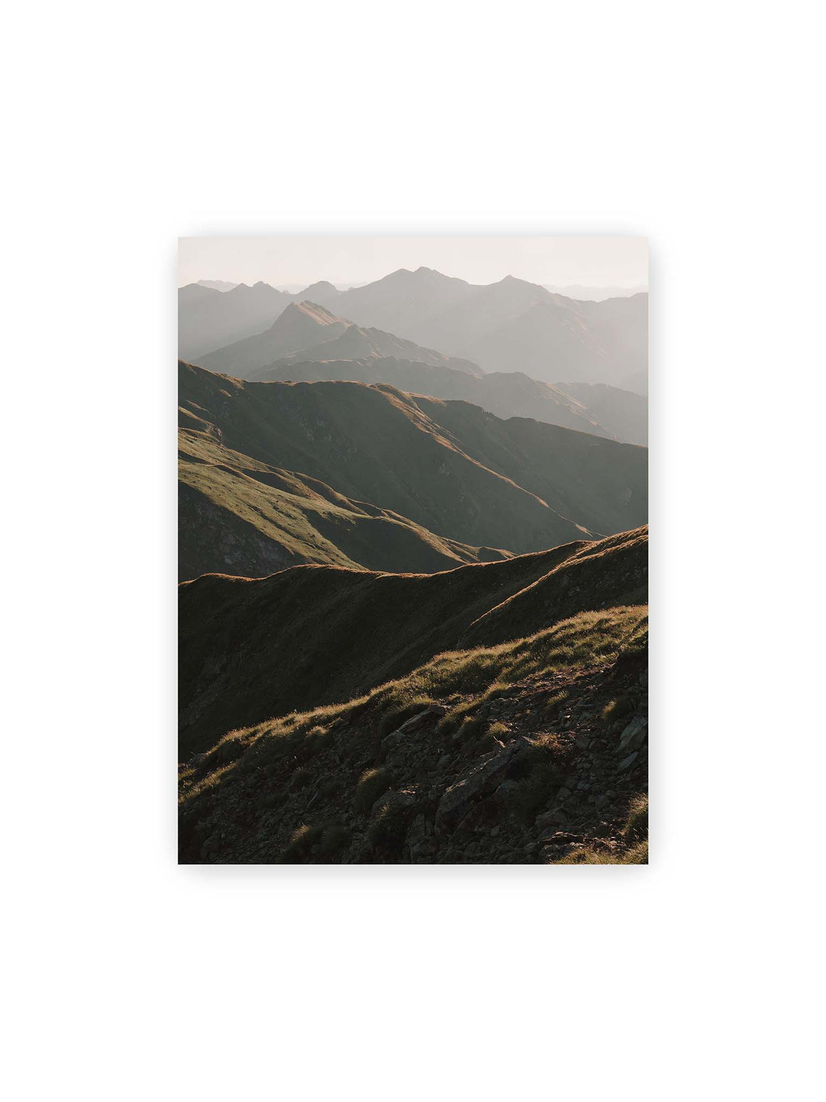 30x40 cm – Gestaffelte Bergketten mit grasbewachsenen Hängen und warm beleuchteten Konturen unter hellem Himmel.