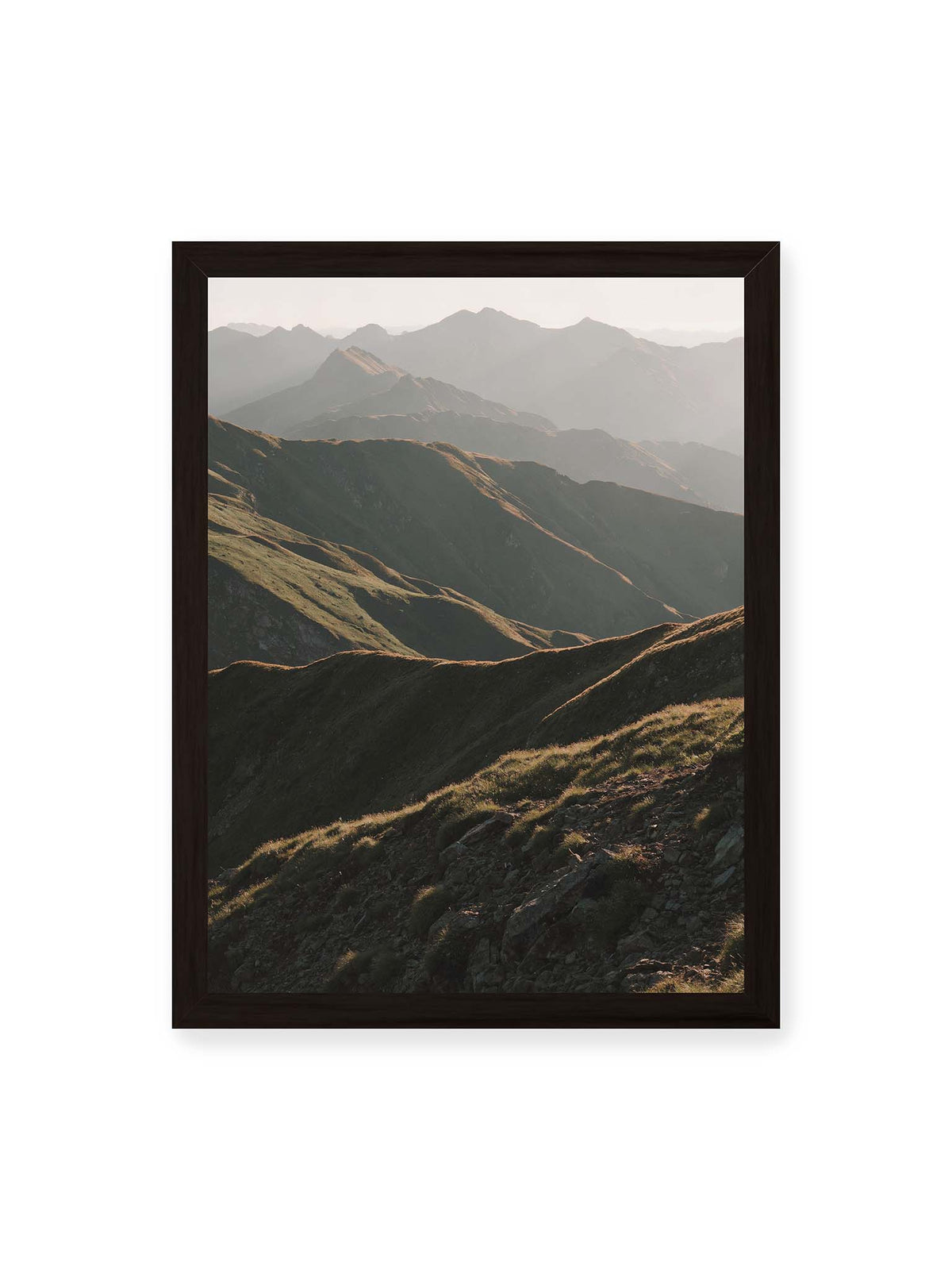 30x40 cm, schwarzer Rahmen – Gestaffelte Bergketten mit grasbewachsenen Hängen und warm beleuchteten Konturen unter hellem Himmel.