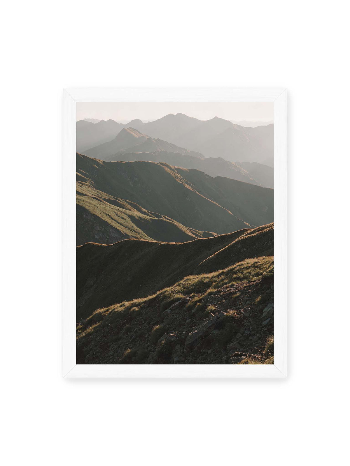 30x40 cm, weißer Rahmen – Gestaffelte Bergketten mit grasbewachsenen Hängen und warm beleuchteten Konturen unter hellem Himmel.