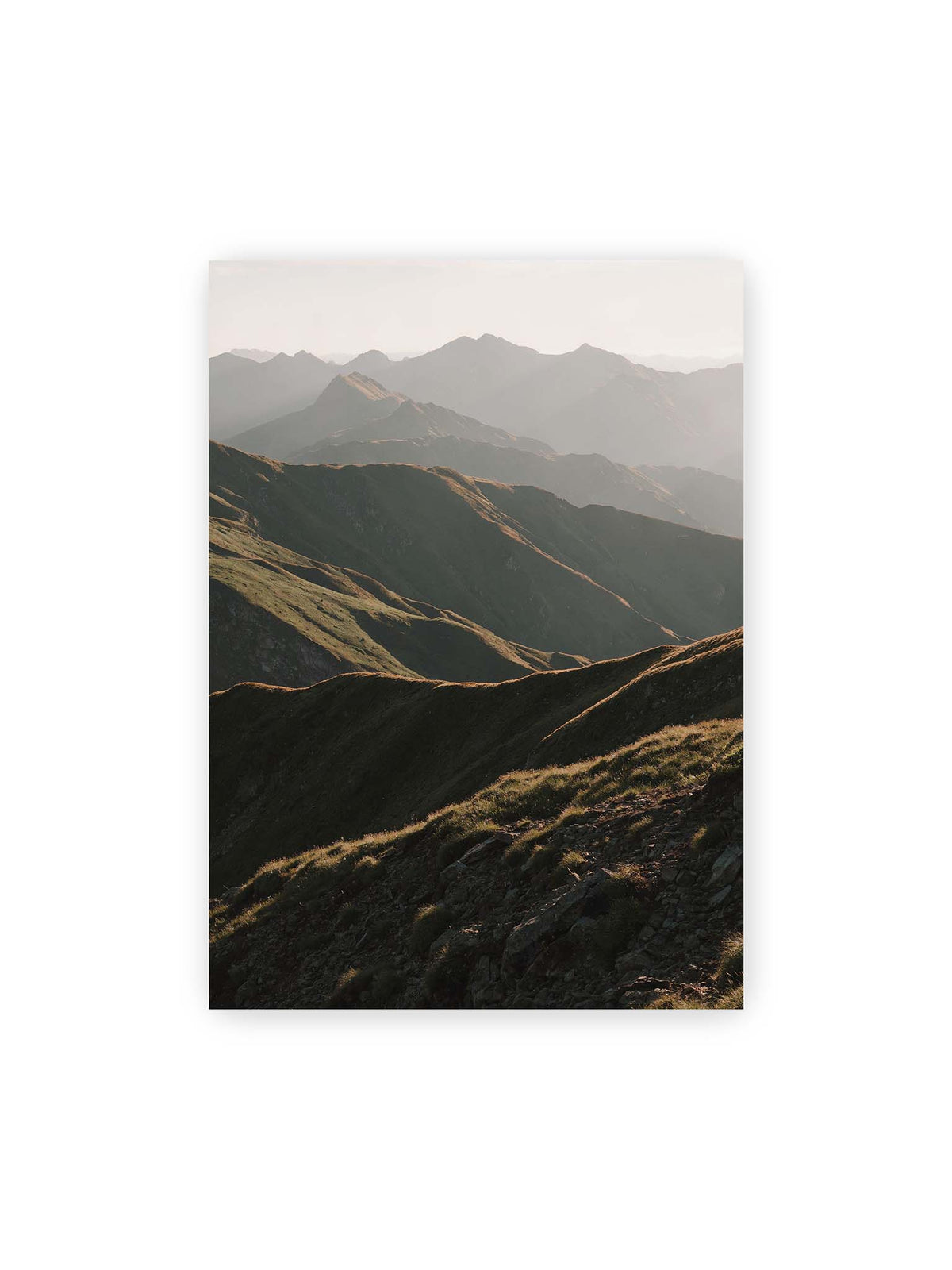 50x70 cm – Gestaffelte Bergketten mit grasbewachsenen Hängen und warm beleuchteten Konturen unter hellem Himmel.