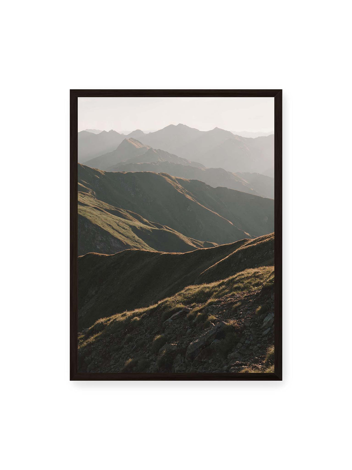 50x70 cm, schwarzer Rahmen – Gestaffelte Bergketten mit grasbewachsenen Hängen und warm beleuchteten Konturen unter hellem Himmel.