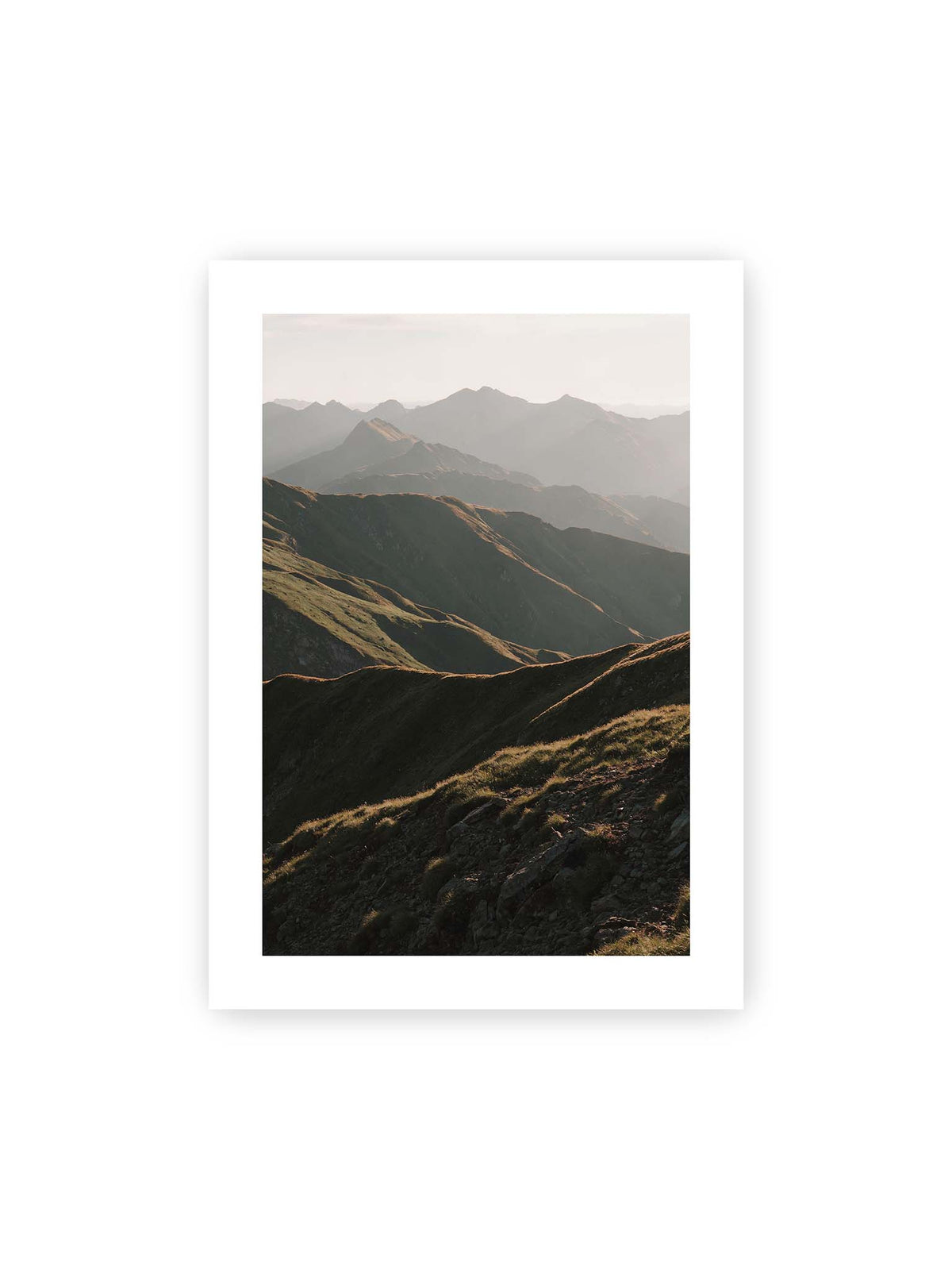 50x70 cm, weißer Galerie-Rand – Gestaffelte Bergketten mit grasbewachsenen Hängen und warm beleuchteten Konturen unter hellem Himmel.