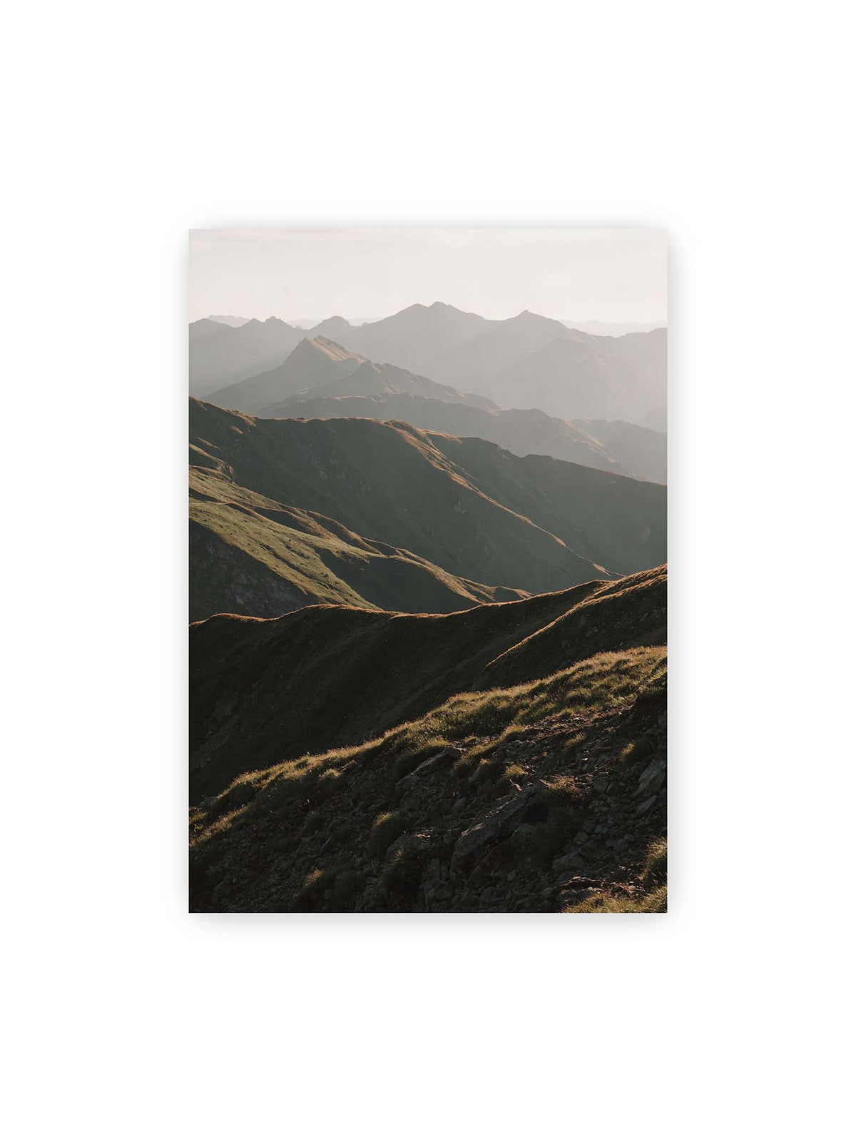 70x100 cm – Gestaffelte Bergketten mit grasbewachsenen Hängen und warm beleuchteten Konturen unter hellem Himmel.