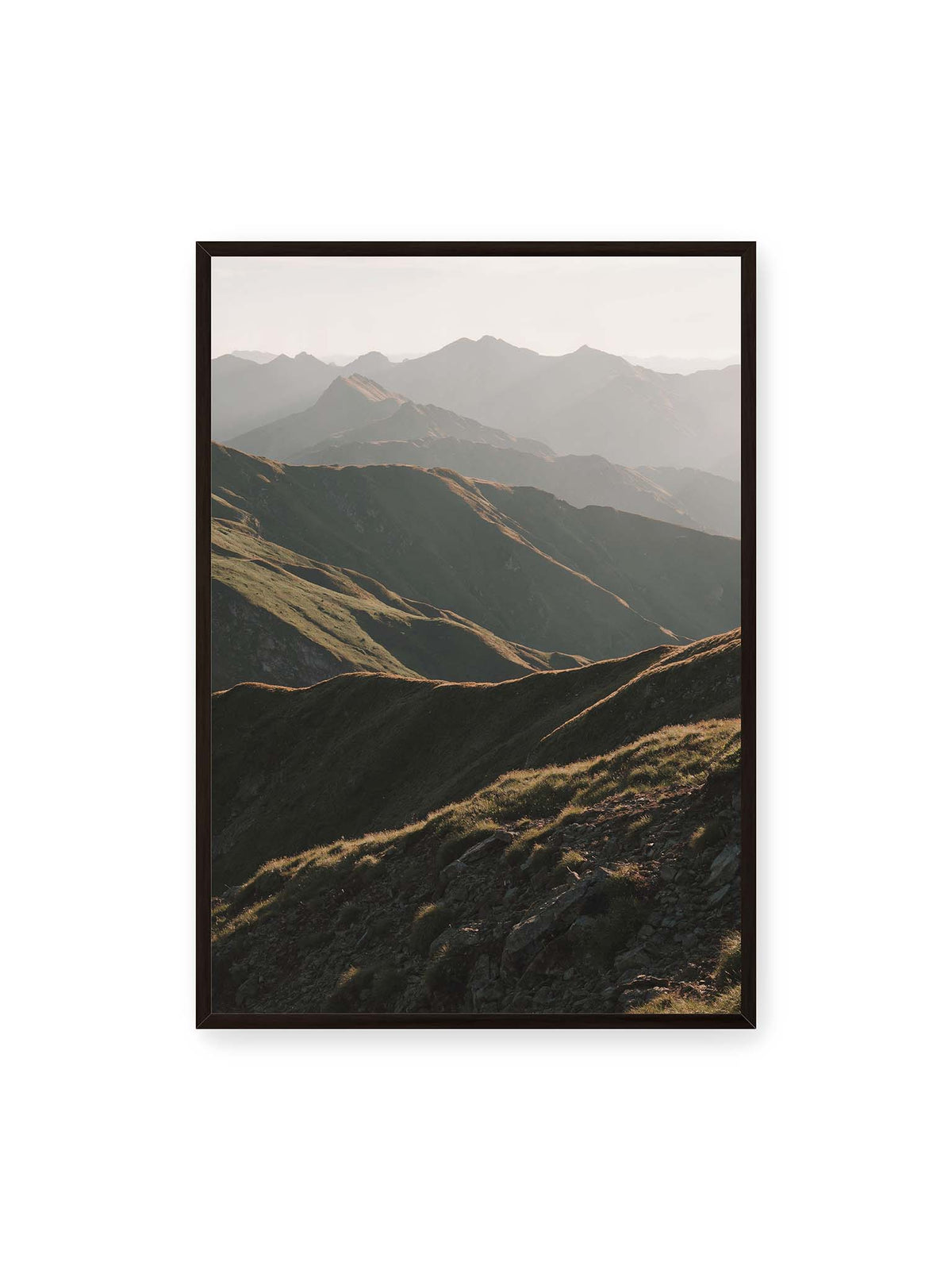 70x100 cm, schwarzer Rahmen – Gestaffelte Bergketten mit grasbewachsenen Hängen und warm beleuchteten Konturen unter hellem Himmel.