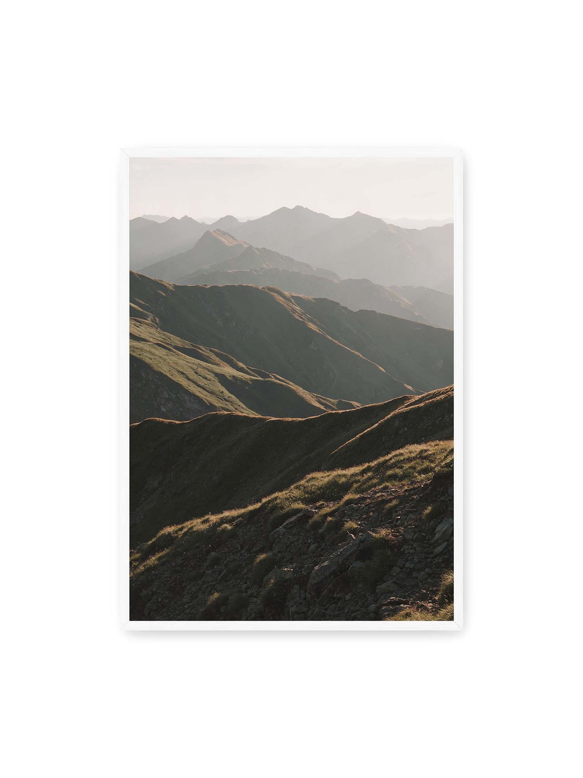 70x100 cm, weißer Rahmen – Gestaffelte Bergketten mit grasbewachsenen Hängen und warm beleuchteten Konturen unter hellem Himmel.