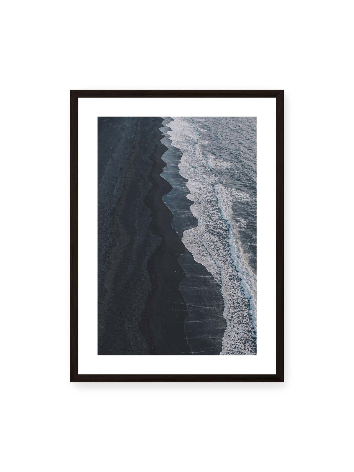 50x70 cm, weißer Galerie-Rand, schwarzer Rahmen – Dunkler Strand aus erhöhter Perspektive mit weißer Brandung und blau-grauem Meer entlang einer geschwungenen Küstenlinie.