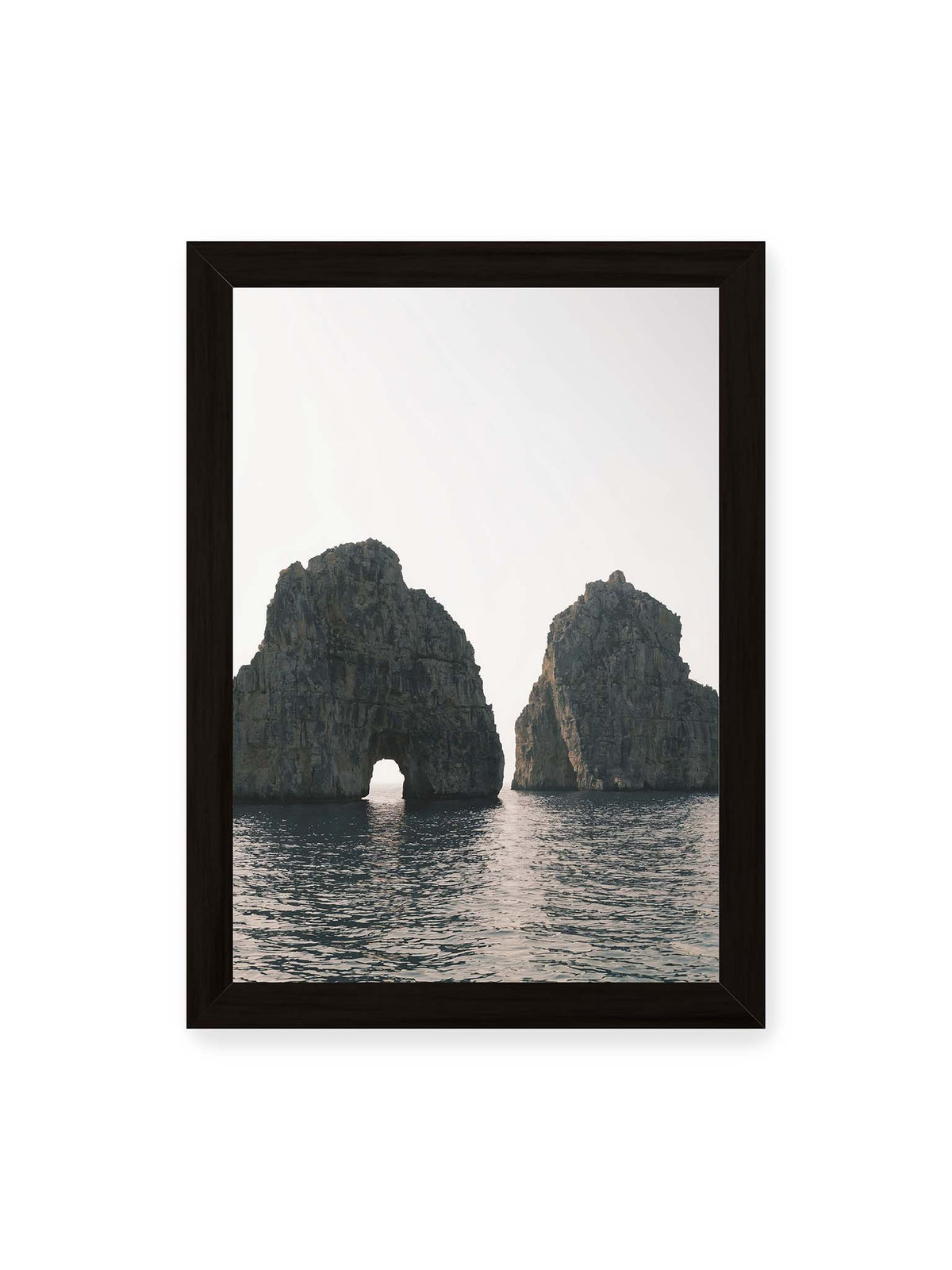 21x30 cm, schwarzer Rahmen – Zwei massive Felsformationen im Meer, eine mit natürlichem Felsbogen, vor hellem Himmel.