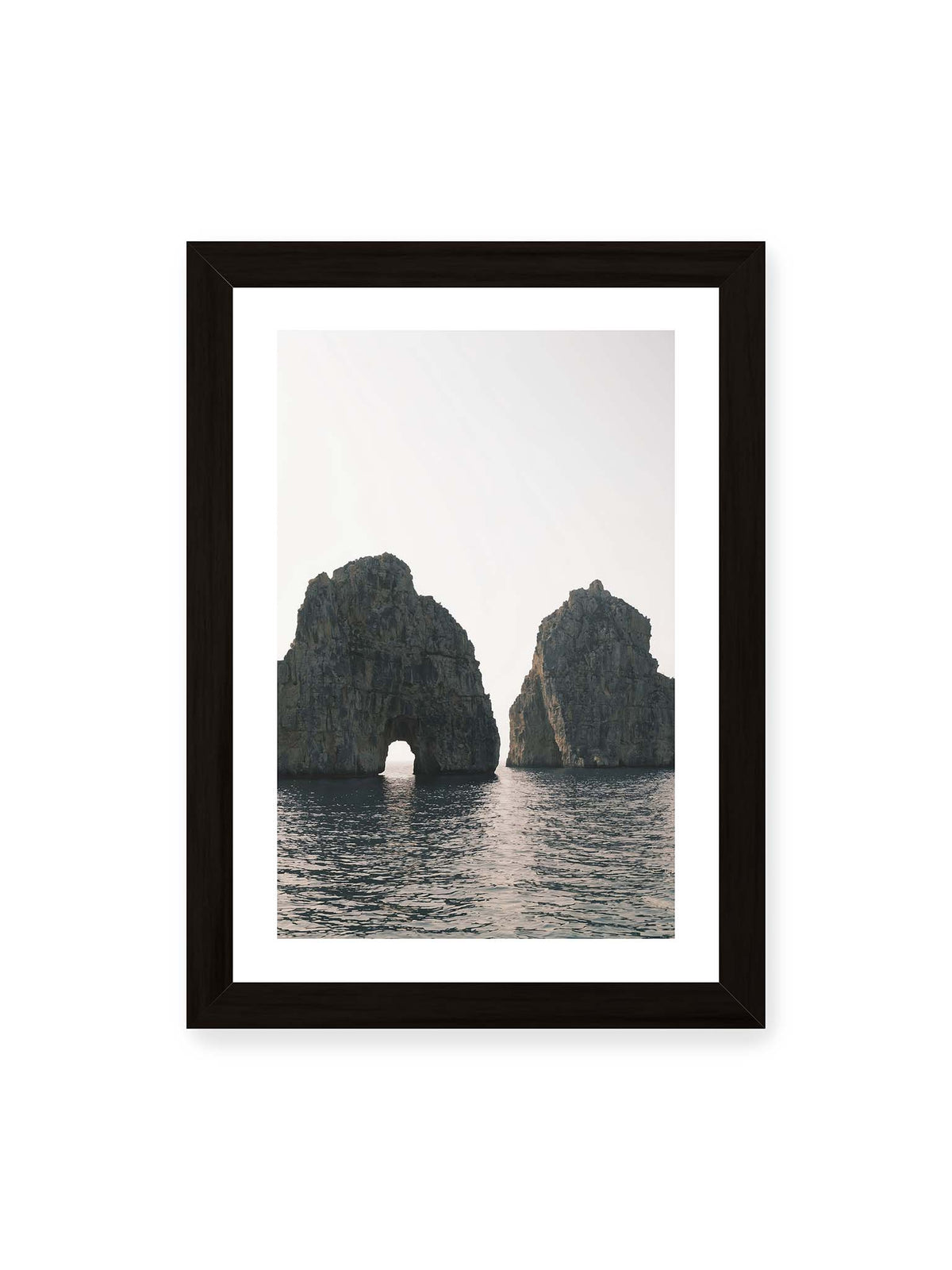 21x30 cm, weißer Galerie-Rand, schwarzer Rahmen – Zwei massive Felsformationen im Meer, eine mit natürlichem Felsbogen, vor hellem Himmel.