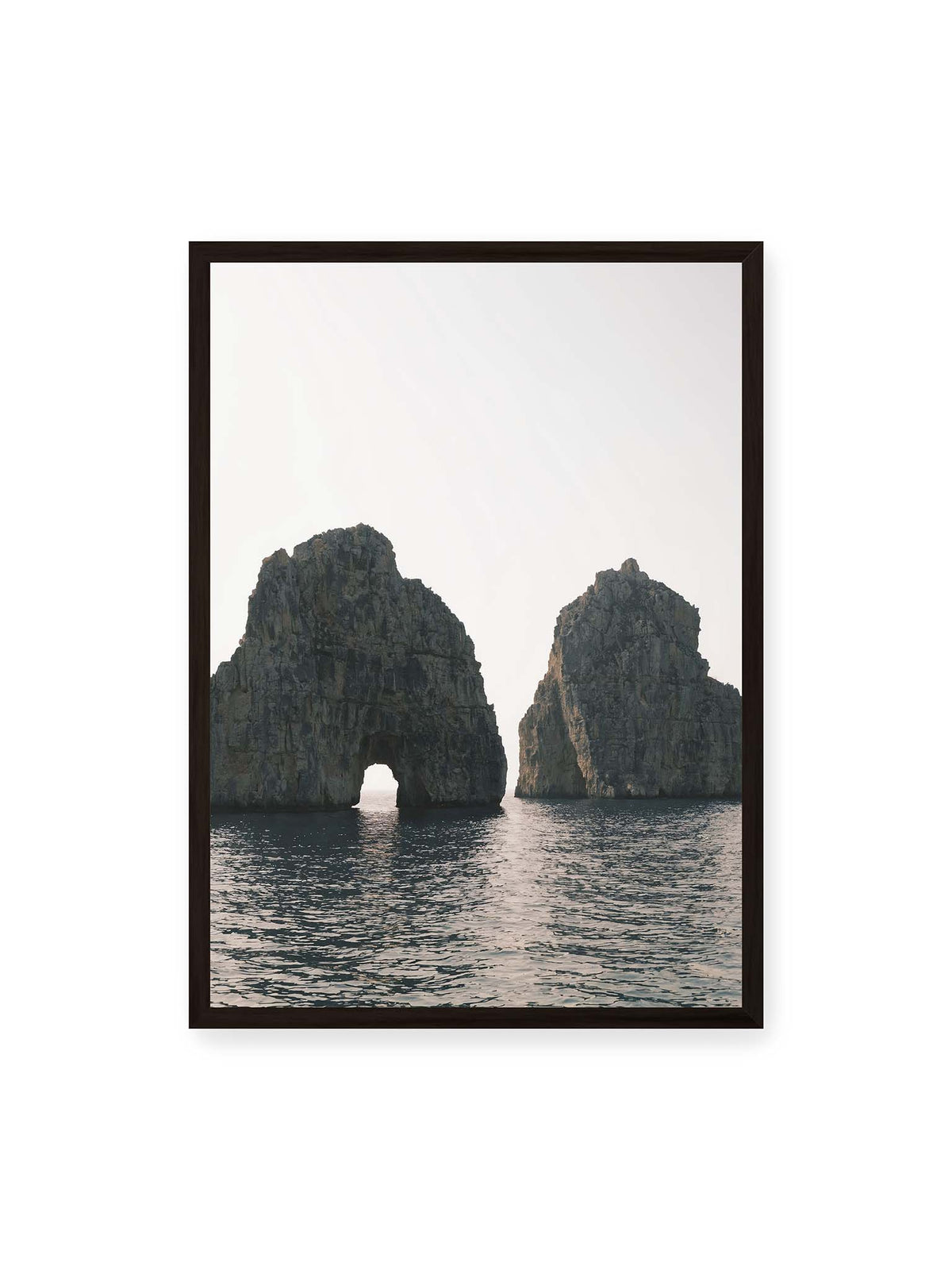 50x70 cm, schwarzer Rahmen – Zwei massive Felsformationen im Meer, eine mit natürlichem Felsbogen, vor hellem Himmel.