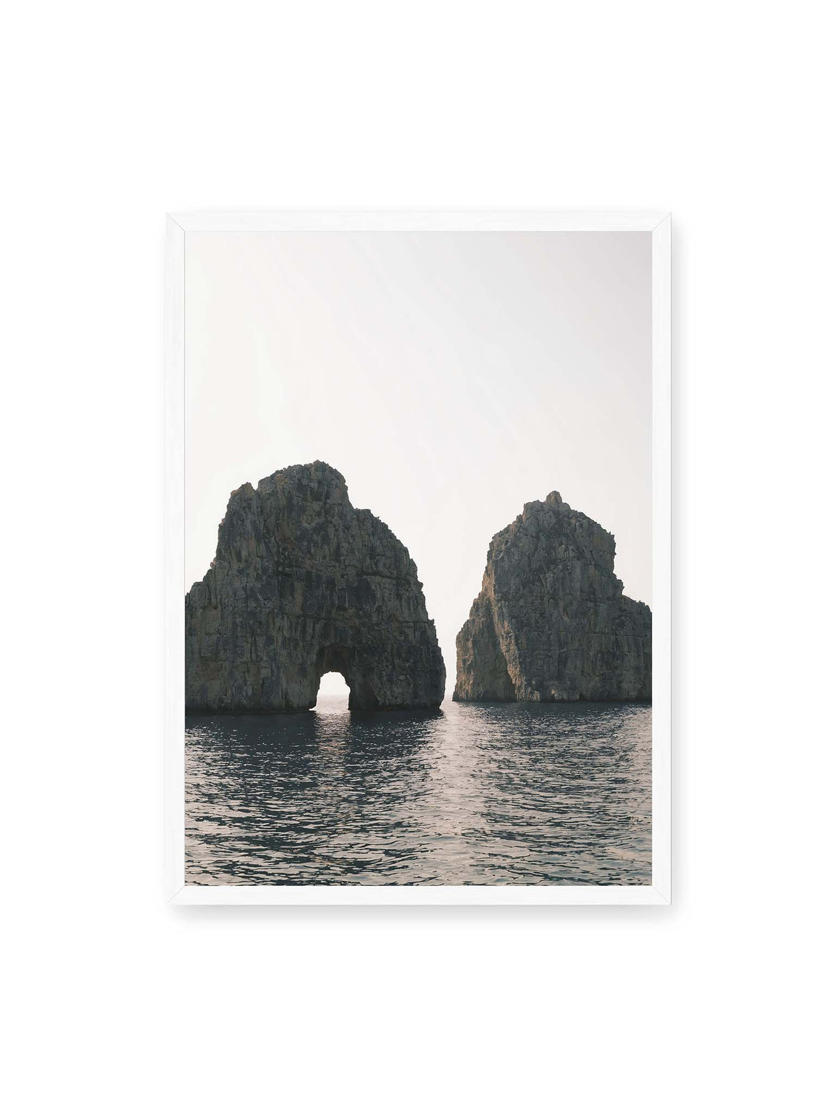 50x70 cm, weißer Rahmen – Zwei massive Felsformationen im Meer, eine mit natürlichem Felsbogen, vor hellem Himmel.