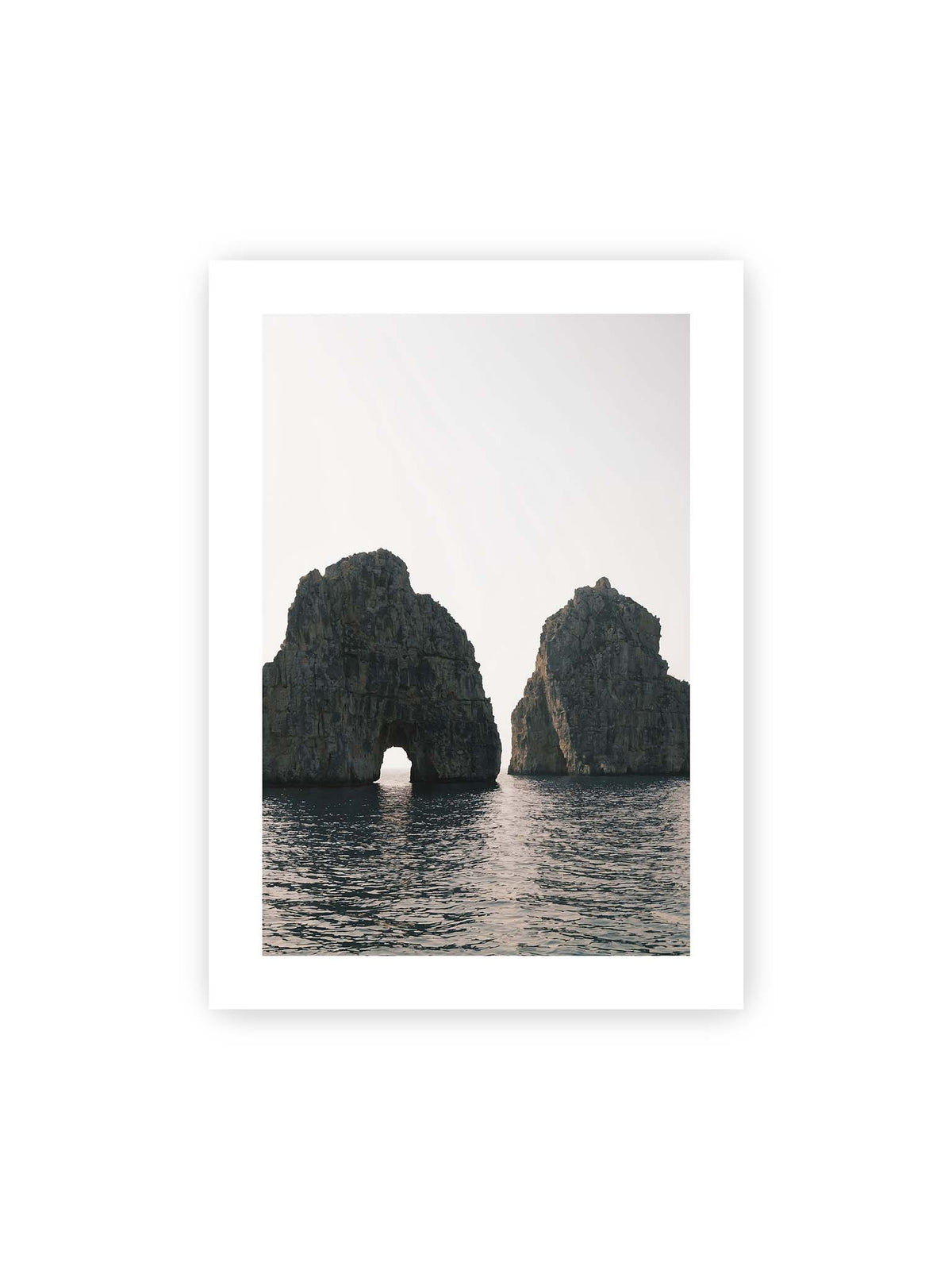 50x70 cm, weißer Galerie-Rand – Zwei massive Felsformationen im Meer, eine mit natürlichem Felsbogen, vor hellem Himmel.