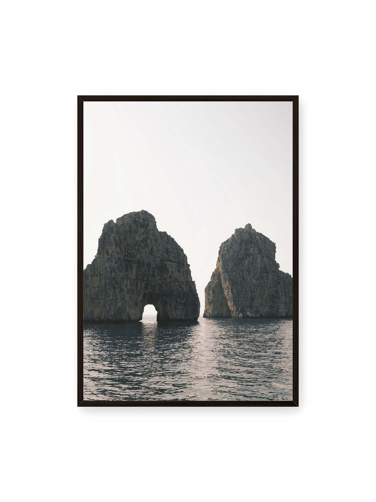 70x100 cm, schwarzer Rahmen – Zwei massive Felsformationen im Meer, eine mit natürlichem Felsbogen, vor hellem Himmel.