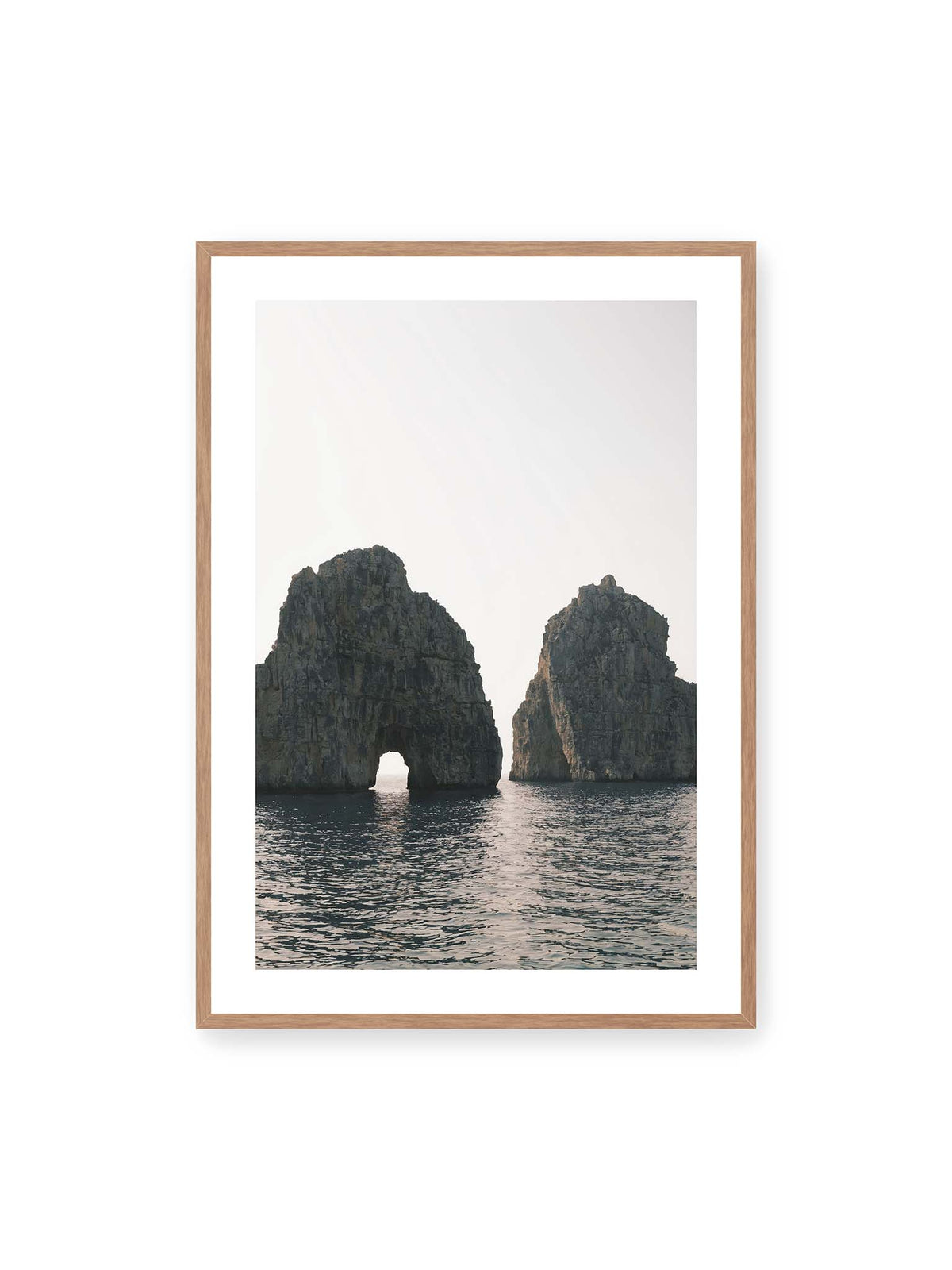 70x100 cm, weißer Galerie-Rand, Eichenrahmen – Zwei massive Felsformationen im Meer, eine mit natürlichem Felsbogen, vor hellem Himmel.
