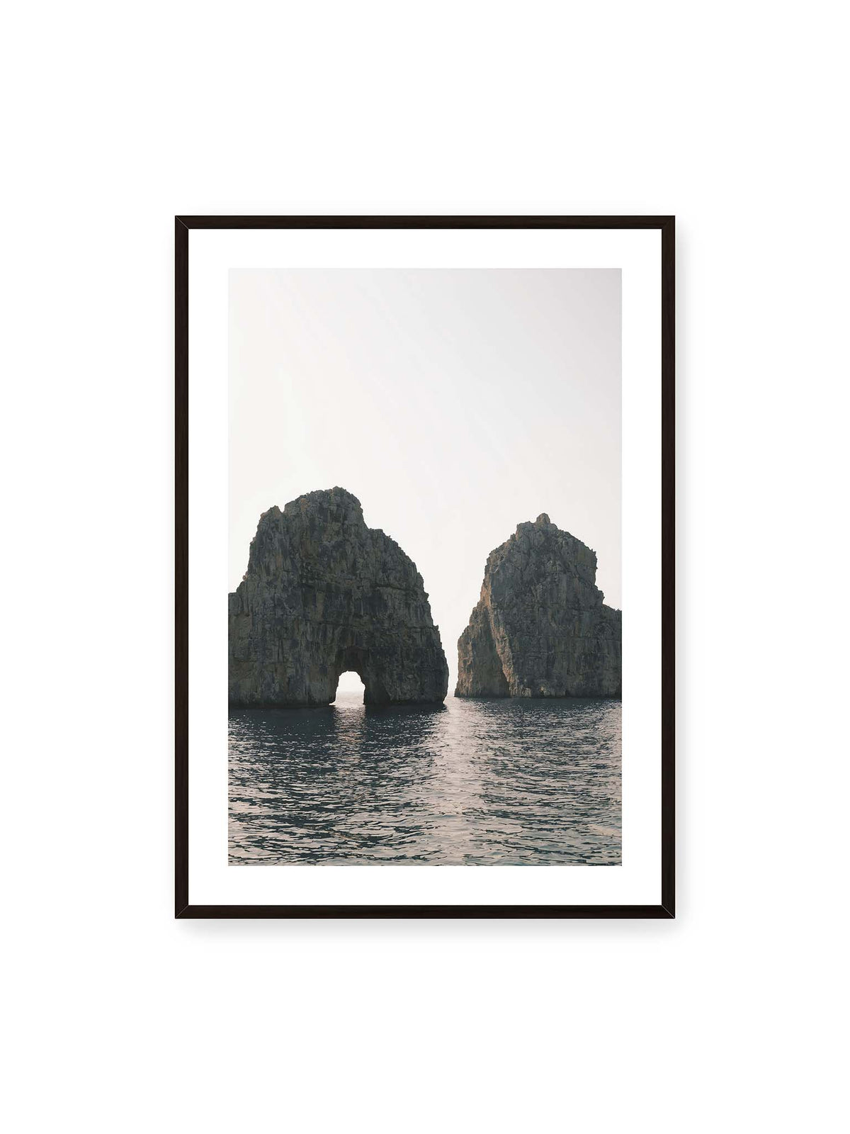 70x100 cm, weißer Galerie-Rand, schwarzer Rahmen – Zwei massive Felsformationen im Meer, eine mit natürlichem Felsbogen, vor hellem Himmel.