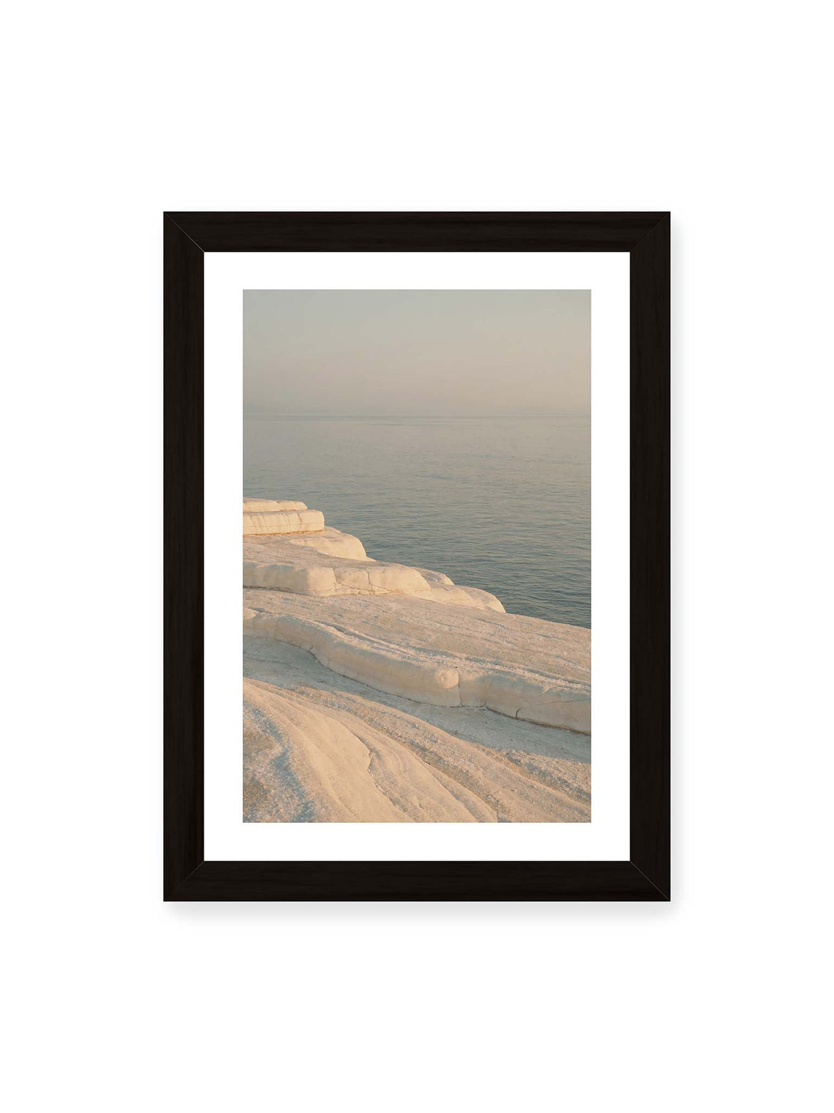 21x30 cm, weißer Galerie-Rand, schwarzer Rahmen – Helle terrassenartige Gesteinsflächen fallen in sanften Stufen zu einem ruhigen, blassblauen Meer ab.
