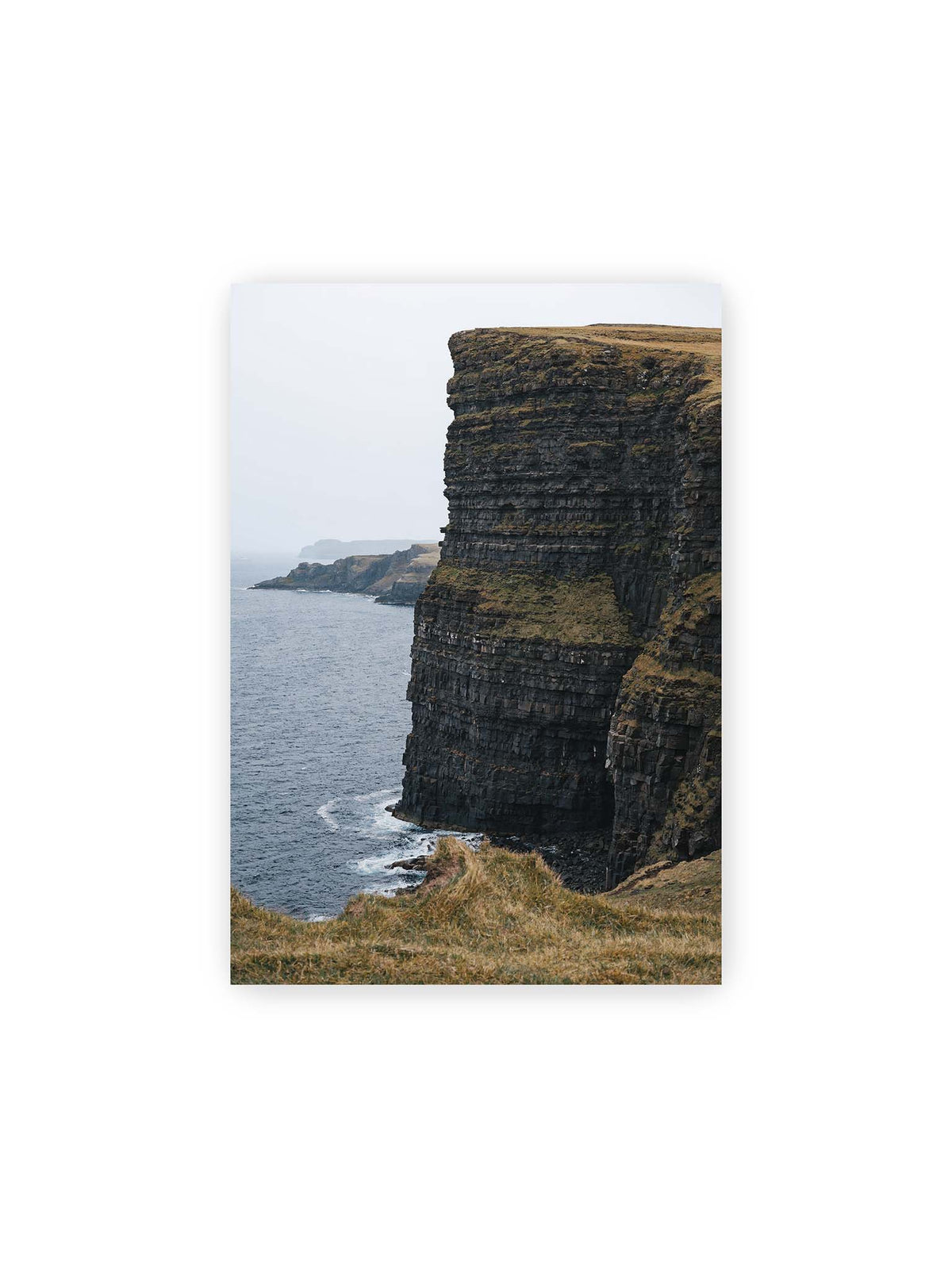 21x30 cm – Dunkle geschichtete Klippen fallen steil zum Meer ab, während weitere Felswände in der hellen Ferne verschwinden.
