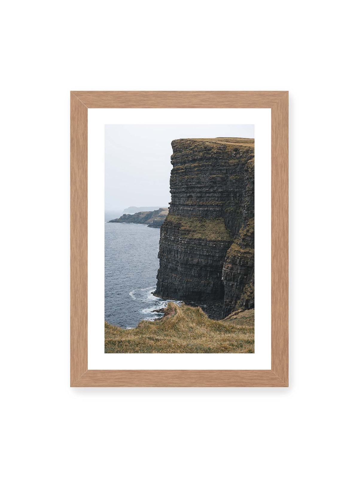 21x30 cm, weißer Galerie-Rand, Eichenrahmen – Dunkle geschichtete Klippen fallen steil zum Meer ab, während weitere Felswände in der hellen Ferne verschwinden.