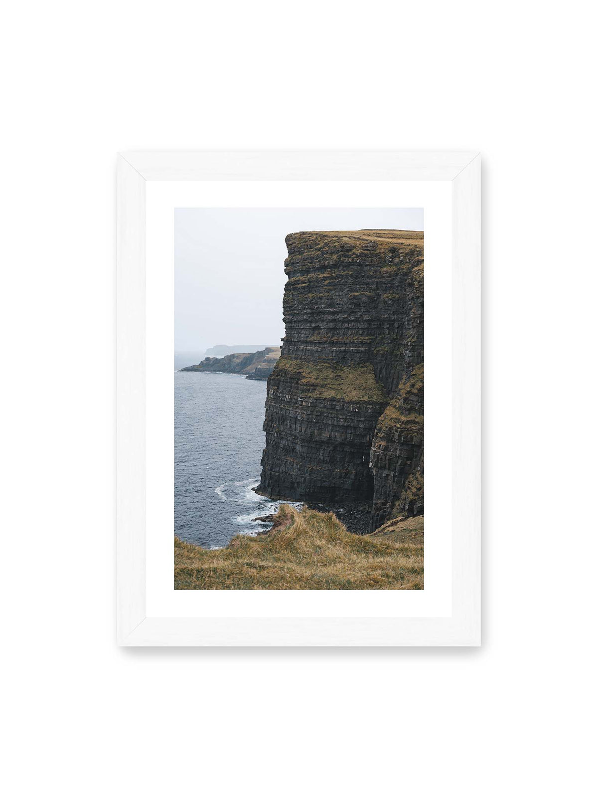 21x30 cm, weißer Galerie-Rand, weißer Rahmen – Dunkle geschichtete Klippen fallen steil zum Meer ab, während weitere Felswände in der hellen Ferne verschwinden.