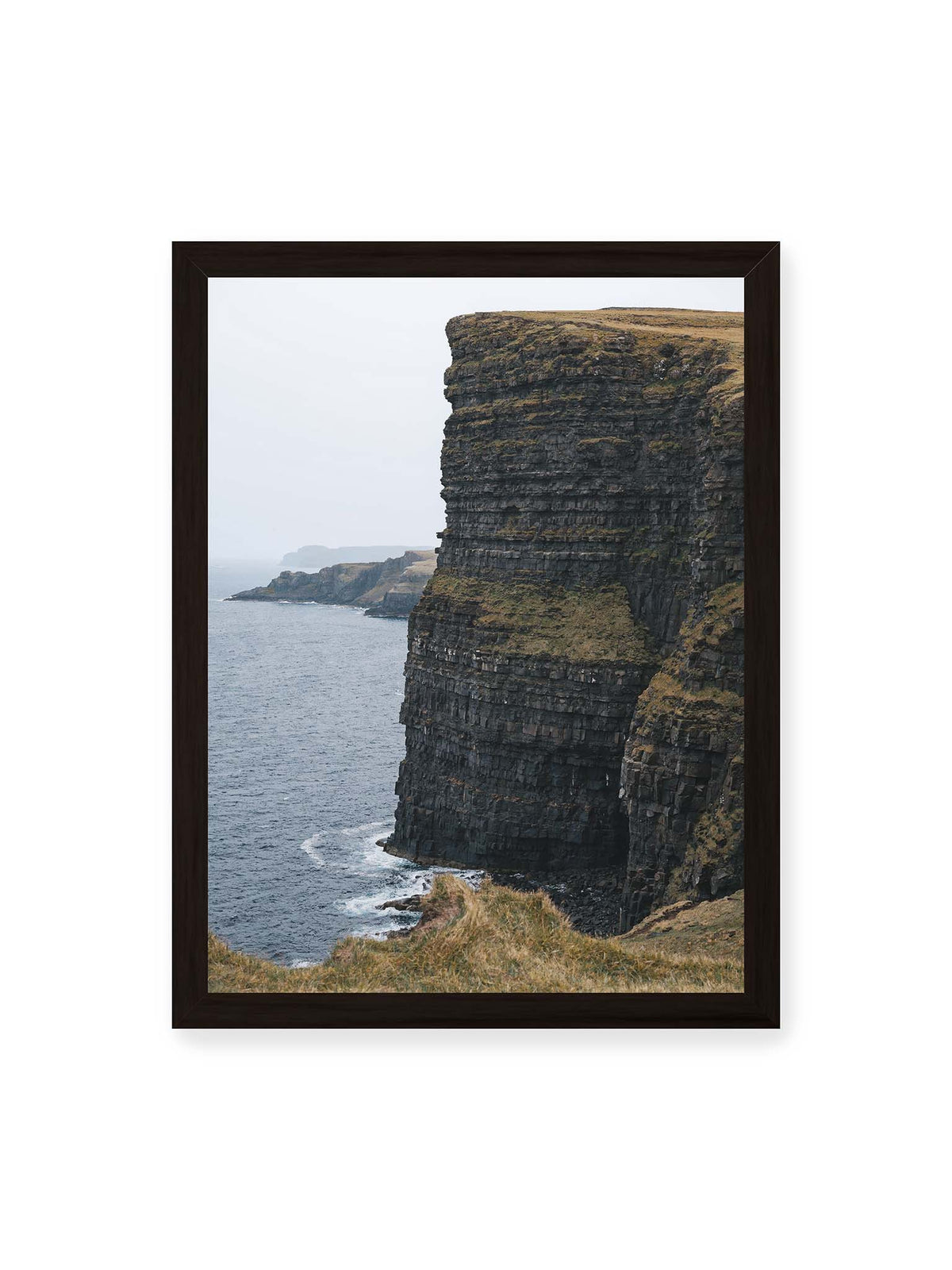 30x40 cm, schwarzer Rahmen – Dunkle geschichtete Klippen fallen steil zum Meer ab, während weitere Felswände in der hellen Ferne verschwinden.