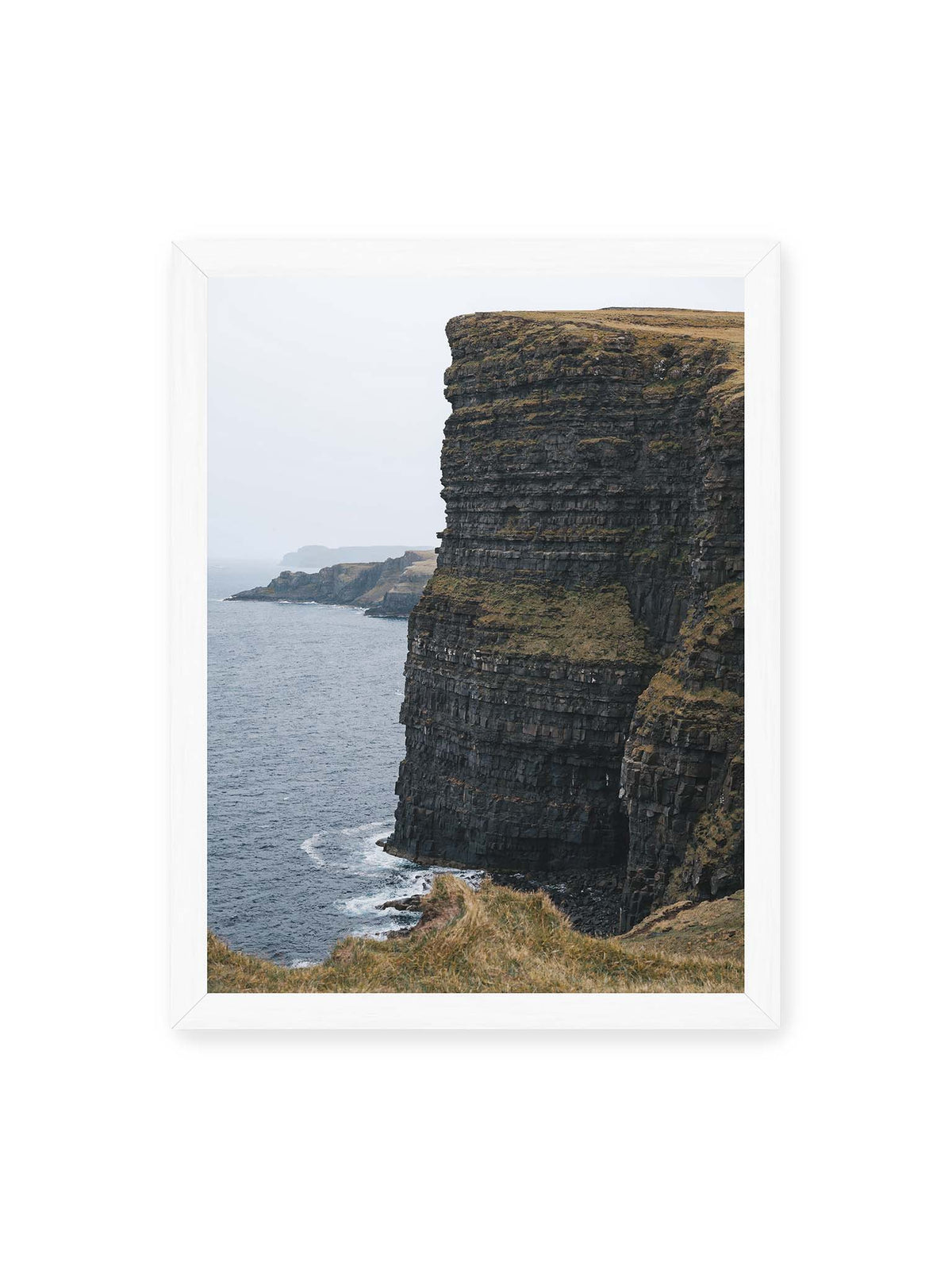 30x40 cm, weißer Rahmen – Dunkle geschichtete Klippen fallen steil zum Meer ab, während weitere Felswände in der hellen Ferne verschwinden.