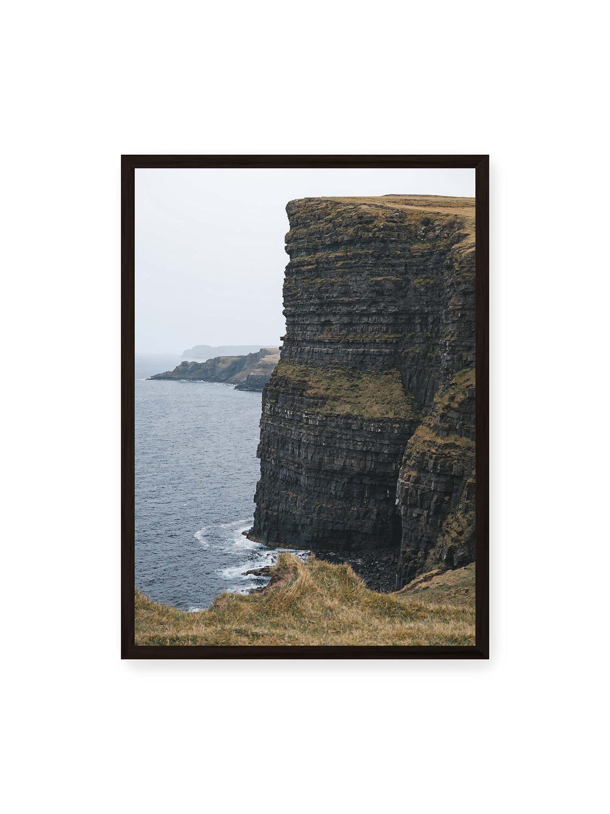 50x70 cm, schwarzer Rahmen – Dunkle geschichtete Klippen fallen steil zum Meer ab, während weitere Felswände in der hellen Ferne verschwinden.