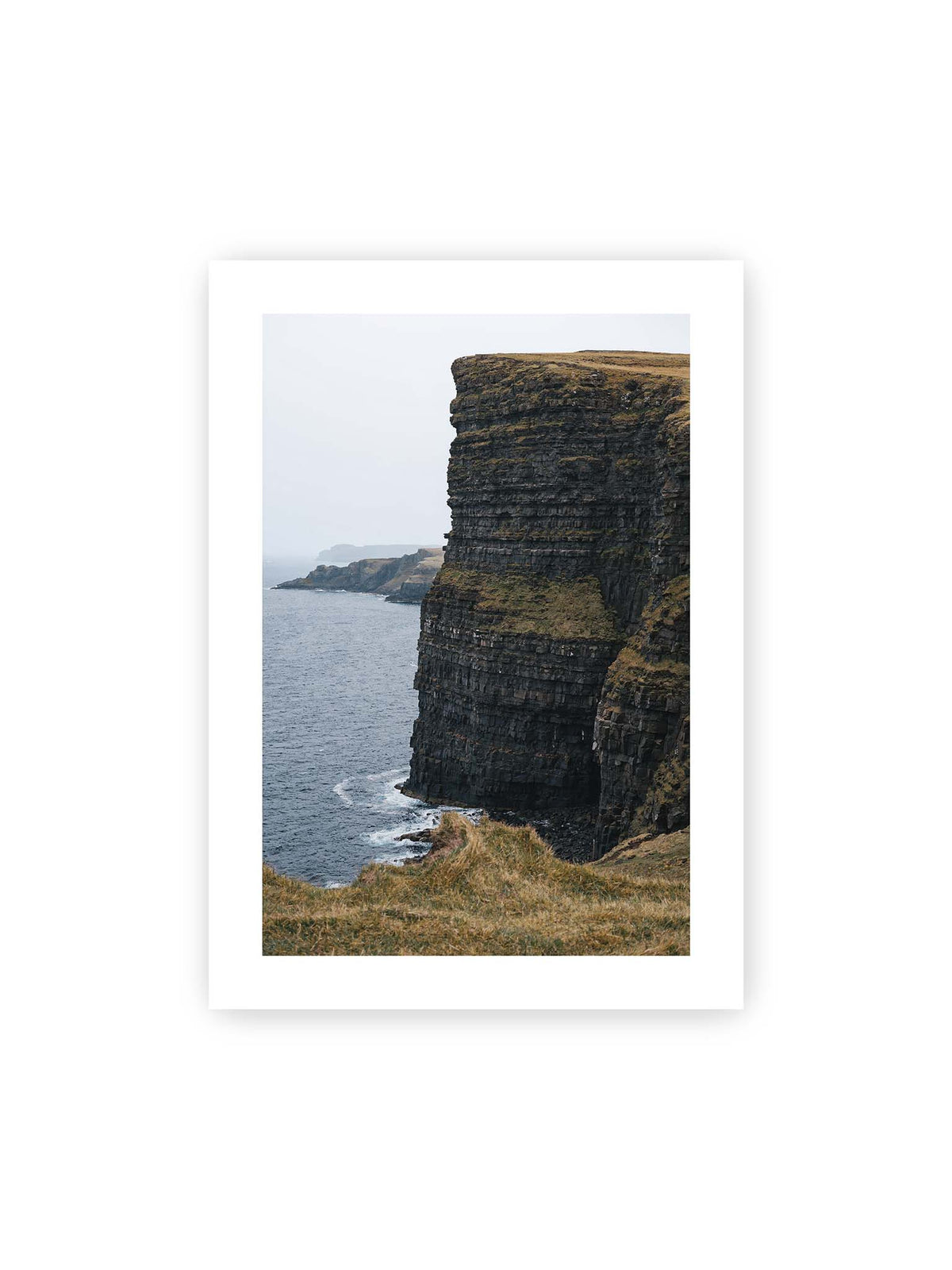 50x70 cm, weißer Galerie-Rand – Dunkle geschichtete Klippen fallen steil zum Meer ab, während weitere Felswände in der hellen Ferne verschwinden.