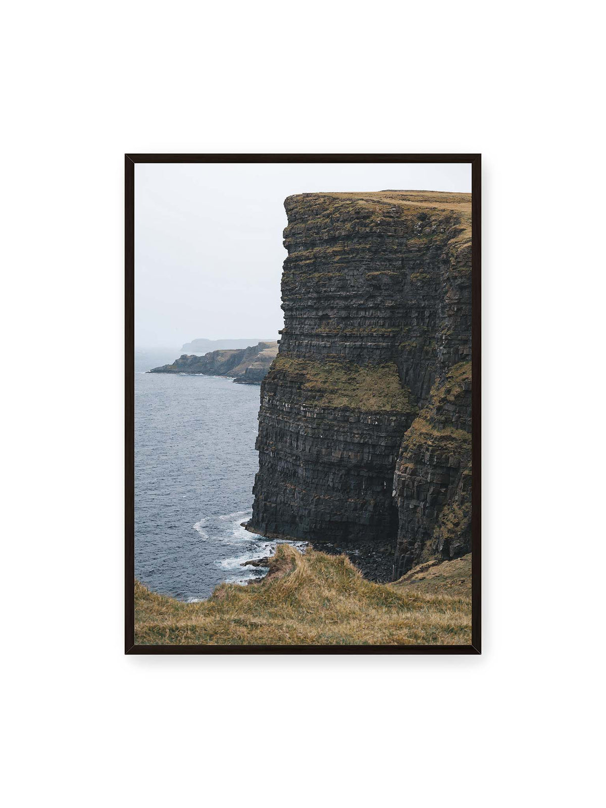 70x100 cm, schwarzer Rahmen – Dunkle geschichtete Klippen fallen steil zum Meer ab, während weitere Felswände in der hellen Ferne verschwinden.