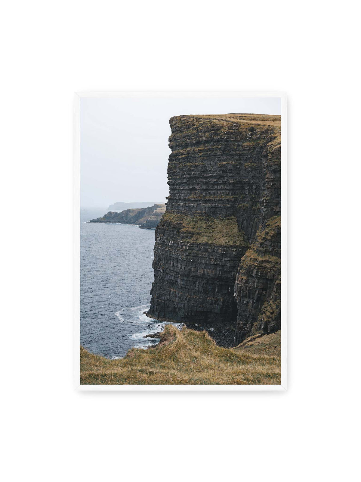 70x100 cm, weißer Rahmen – Dunkle geschichtete Klippen fallen steil zum Meer ab, während weitere Felswände in der hellen Ferne verschwinden.