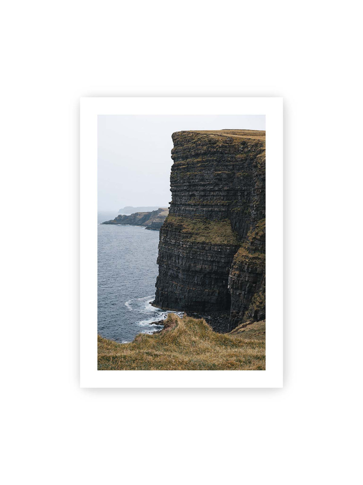70x100 cm, weißer Galerie-Rand – Dunkle geschichtete Klippen fallen steil zum Meer ab, während weitere Felswände in der hellen Ferne verschwinden.