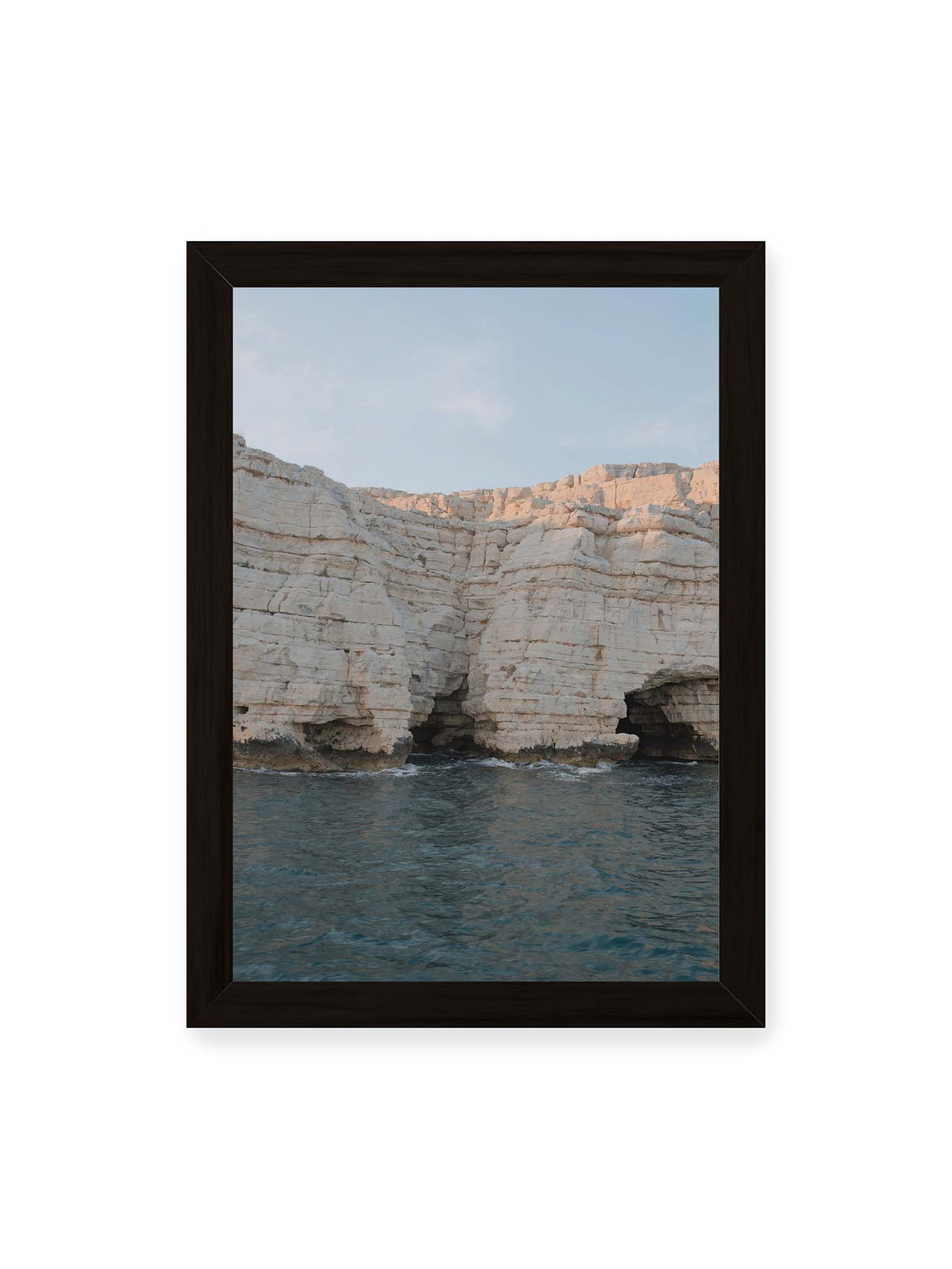 21x30 cm, schwarzer Rahmen – Helle geschichtete Felswand mit dunklen Grotten über bewegtem blauem Meer und blassem Himmel.