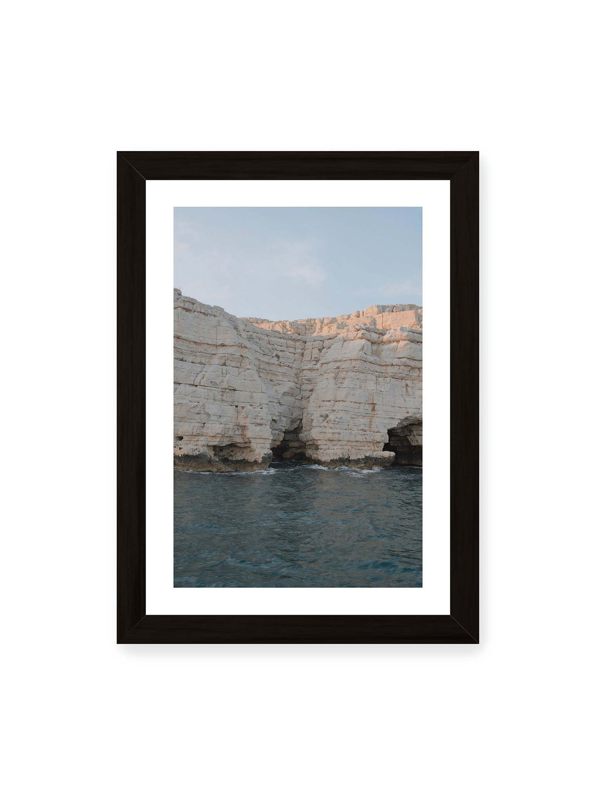 21x30 cm, weißer Galerie-Rand, schwarzer Rahmen – Helle geschichtete Felswand mit dunklen Grotten über bewegtem blauem Meer und blassem Himmel.