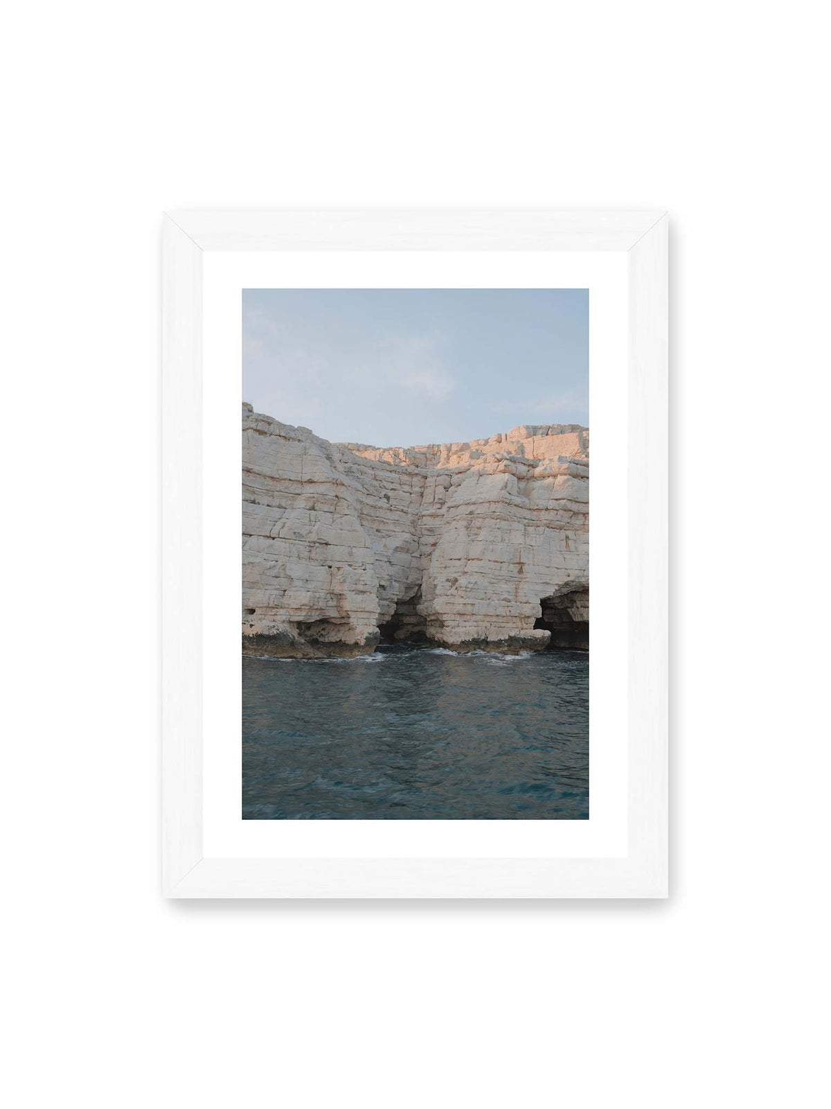 21x30 cm, weißer Galerie-Rand, weißer Rahmen – Helle geschichtete Felswand mit dunklen Grotten über bewegtem blauem Meer und blassem Himmel.