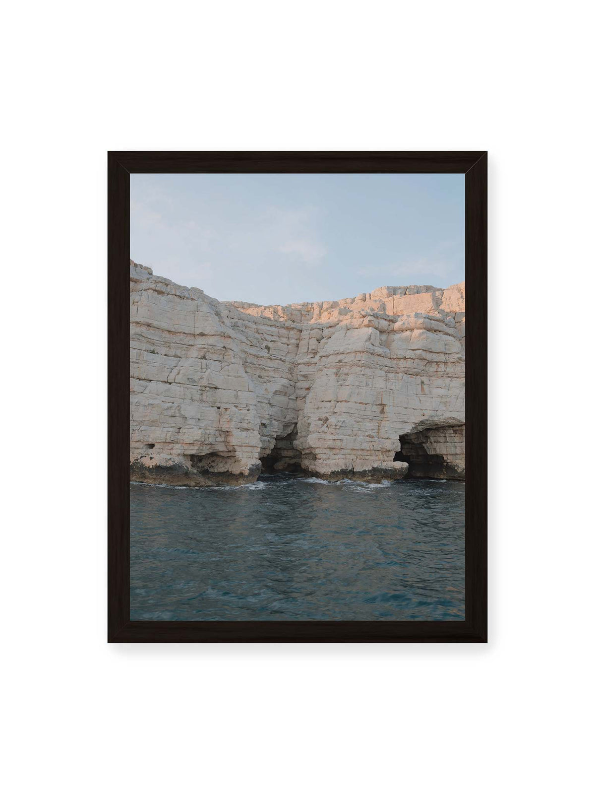 30x40 cm, schwarzer Rahmen – Helle geschichtete Felswand mit dunklen Grotten über bewegtem blauem Meer und blassem Himmel.