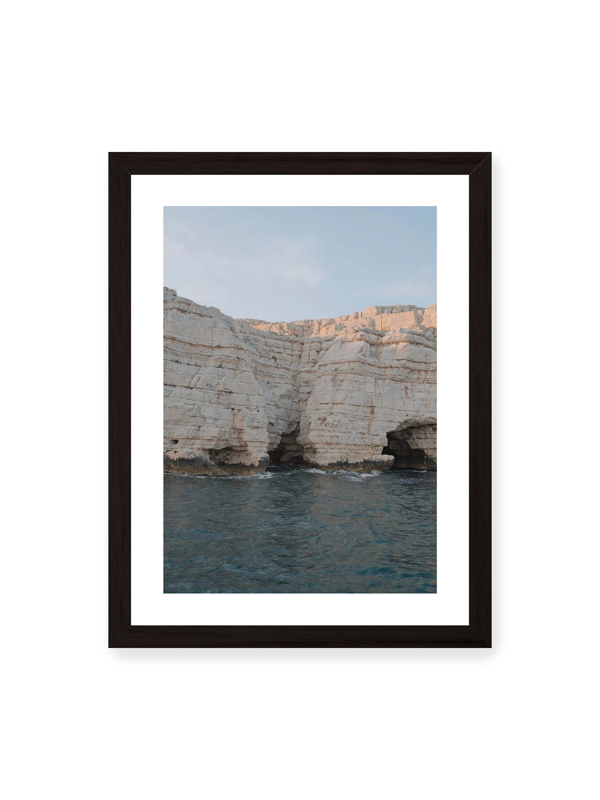 30x40 cm, weißer Galerie-Rand, schwarzer Rahmen – Helle geschichtete Felswand mit dunklen Grotten über bewegtem blauem Meer und blassem Himmel.