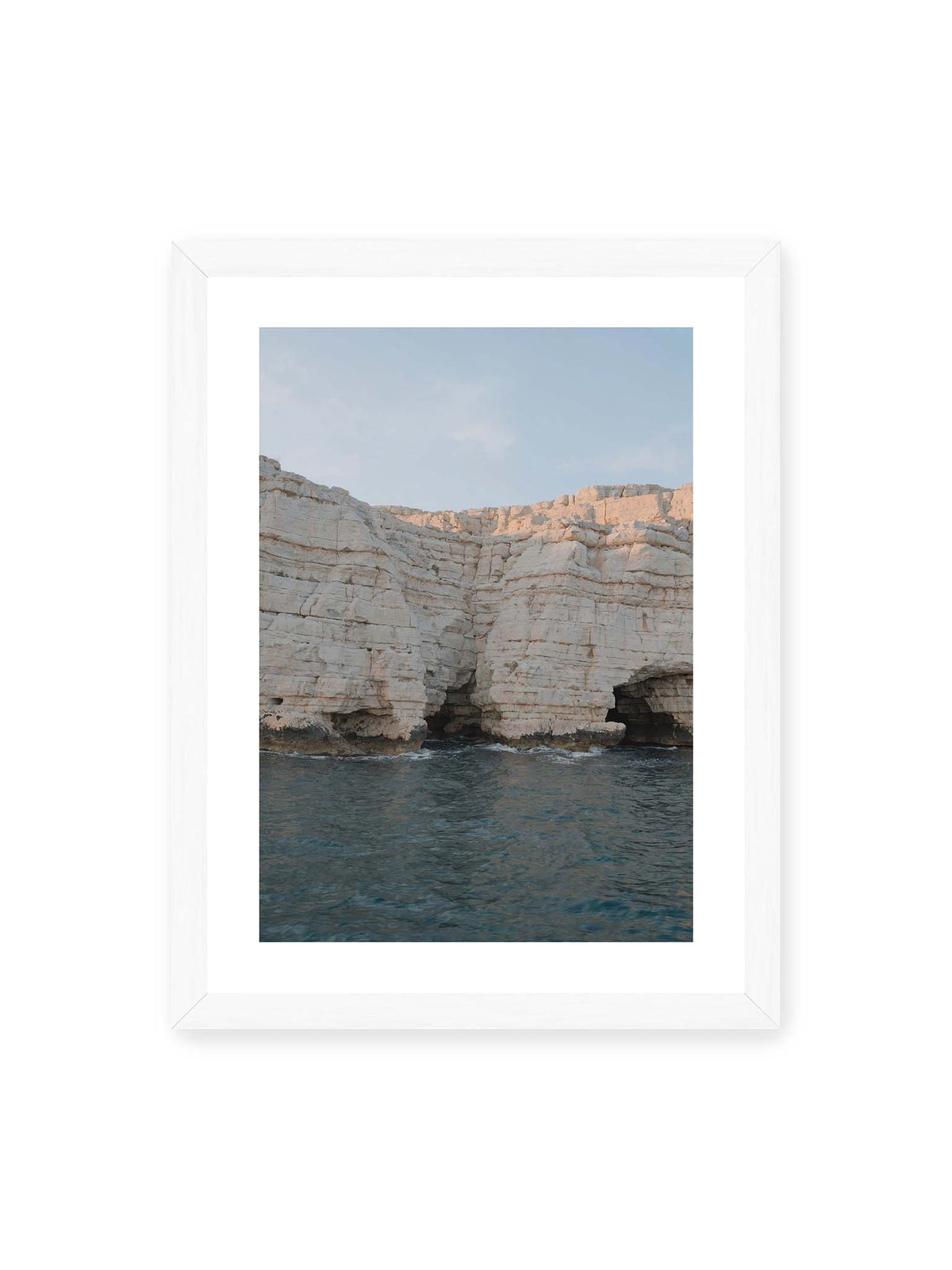30x40 cm, weißer Galerie-Rand, weißer Rahmen – Helle geschichtete Felswand mit dunklen Grotten über bewegtem blauem Meer und blassem Himmel.