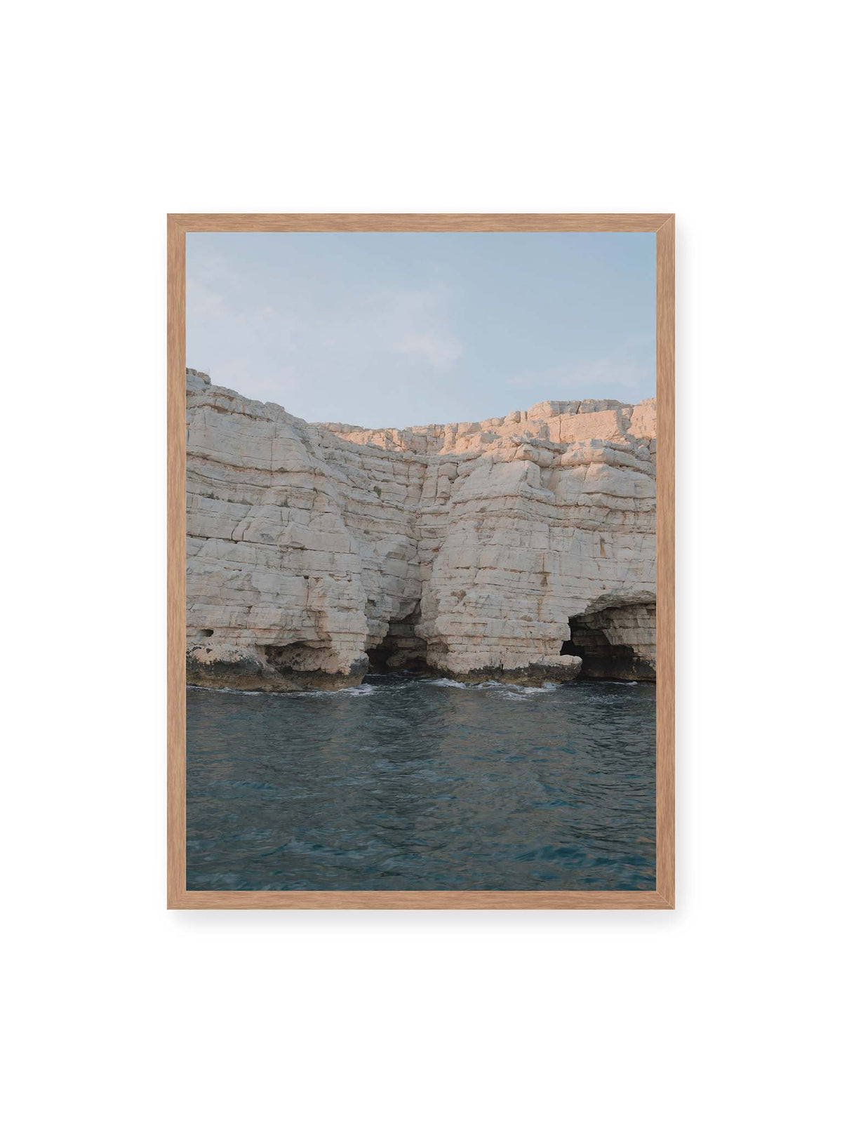 50x70 cm, Eichenrahmen – Helle geschichtete Felswand mit dunklen Grotten über bewegtem blauem Meer und blassem Himmel.