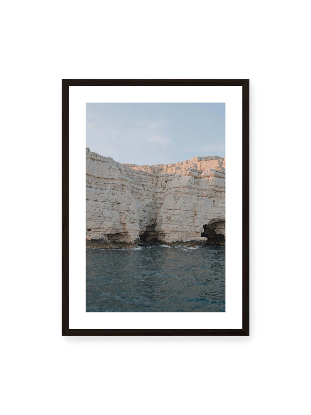 50x70 cm, weißer Galerie-Rand, schwarzer Rahmen – Helle geschichtete Felswand mit dunklen Grotten über bewegtem blauem Meer und blassem Himmel.