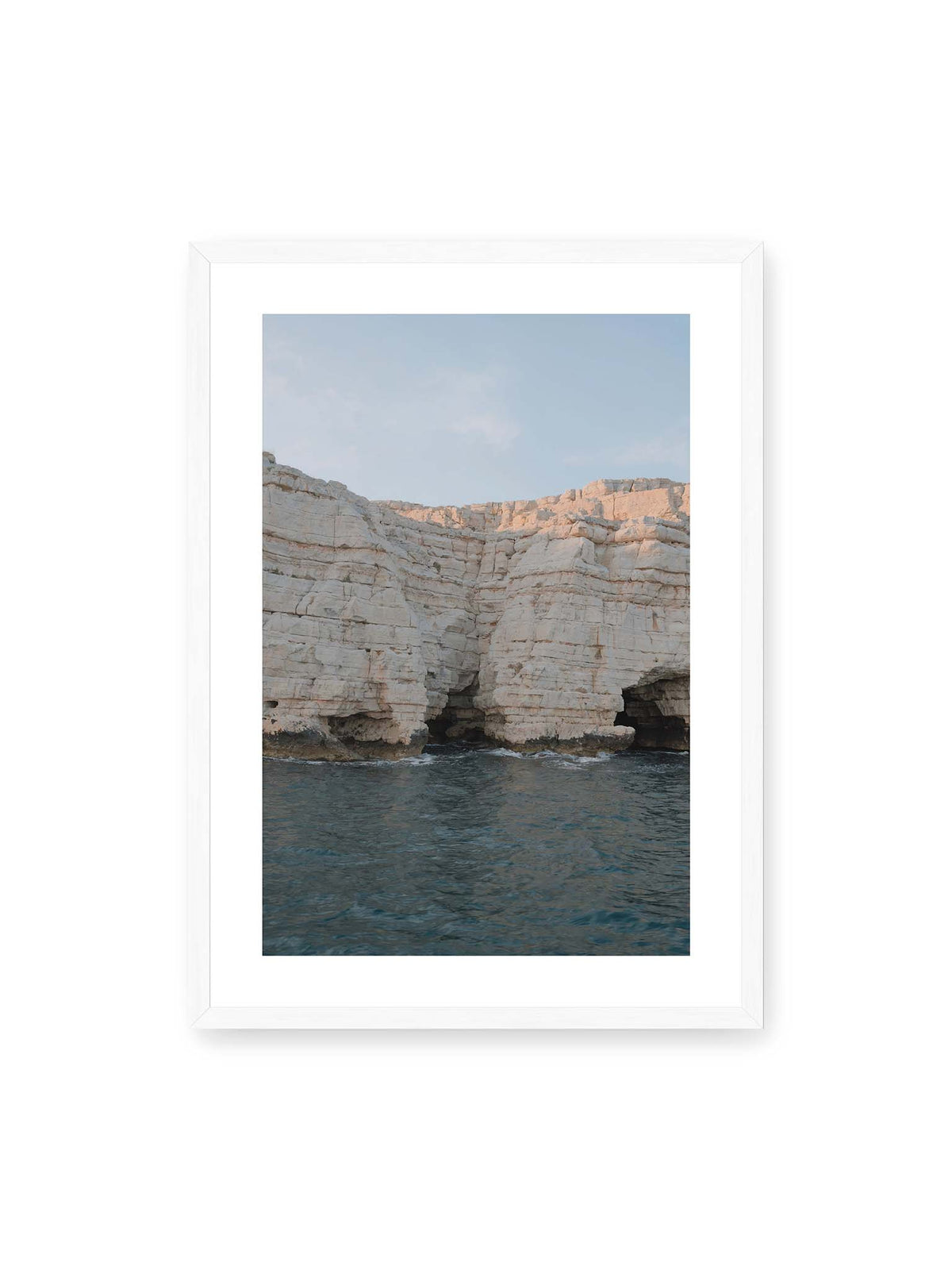 50x70 cm, weißer Galerie-Rand, weißer Rahmen – Helle geschichtete Felswand mit dunklen Grotten über bewegtem blauem Meer und blassem Himmel.