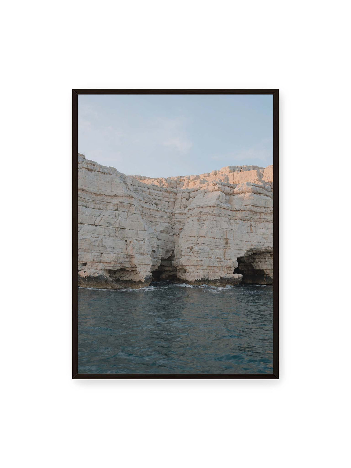 70x100 cm, schwarzer Rahmen – Helle geschichtete Felswand mit dunklen Grotten über bewegtem blauem Meer und blassem Himmel.