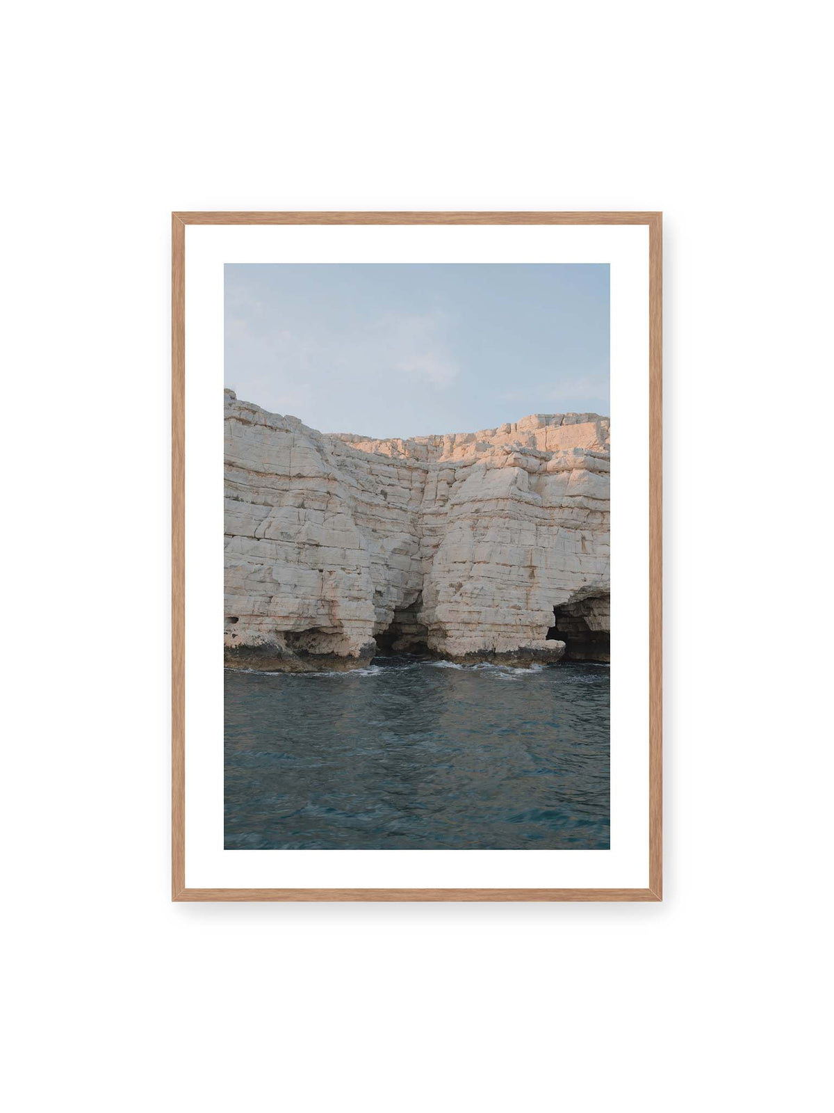 70x100 cm, weißer Galerie-Rand, Eichenrahmen – Helle geschichtete Felswand mit dunklen Grotten über bewegtem blauem Meer und blassem Himmel.