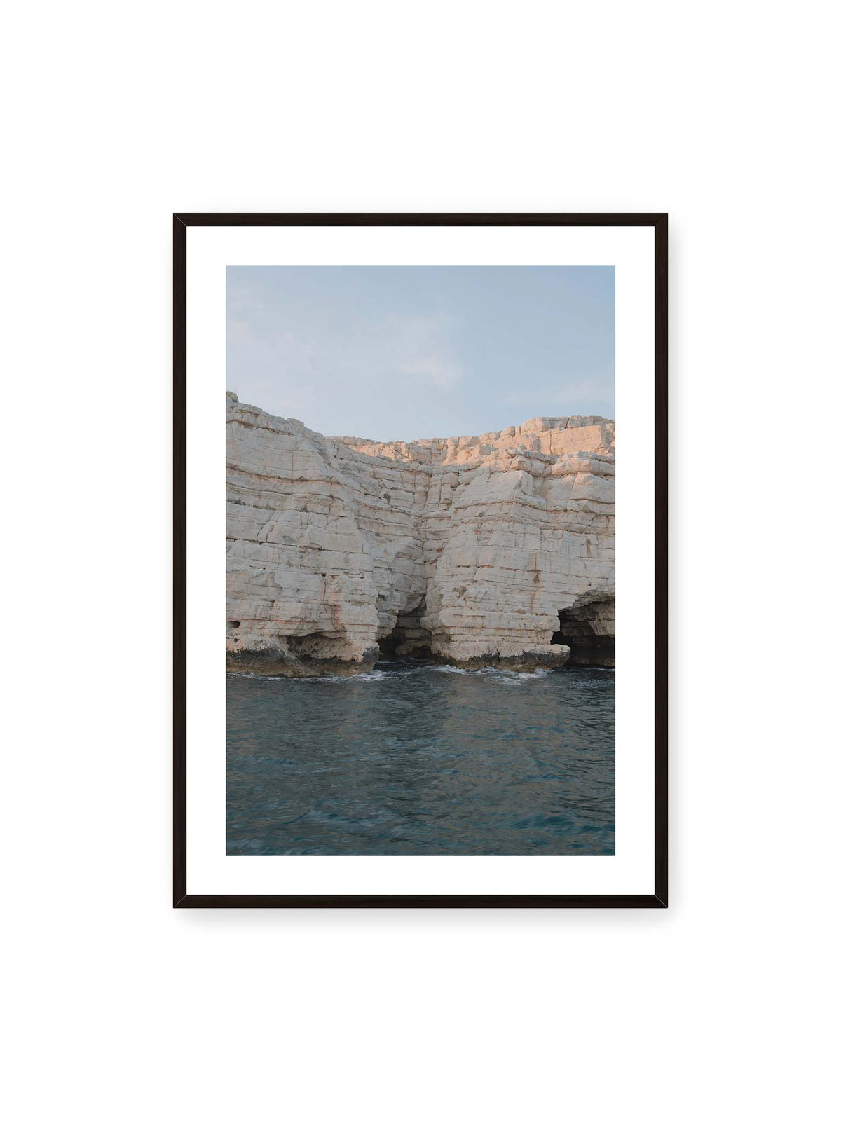 70x100 cm, weißer Galerie-Rand, schwarzer Rahmen – Helle geschichtete Felswand mit dunklen Grotten über bewegtem blauem Meer und blassem Himmel.