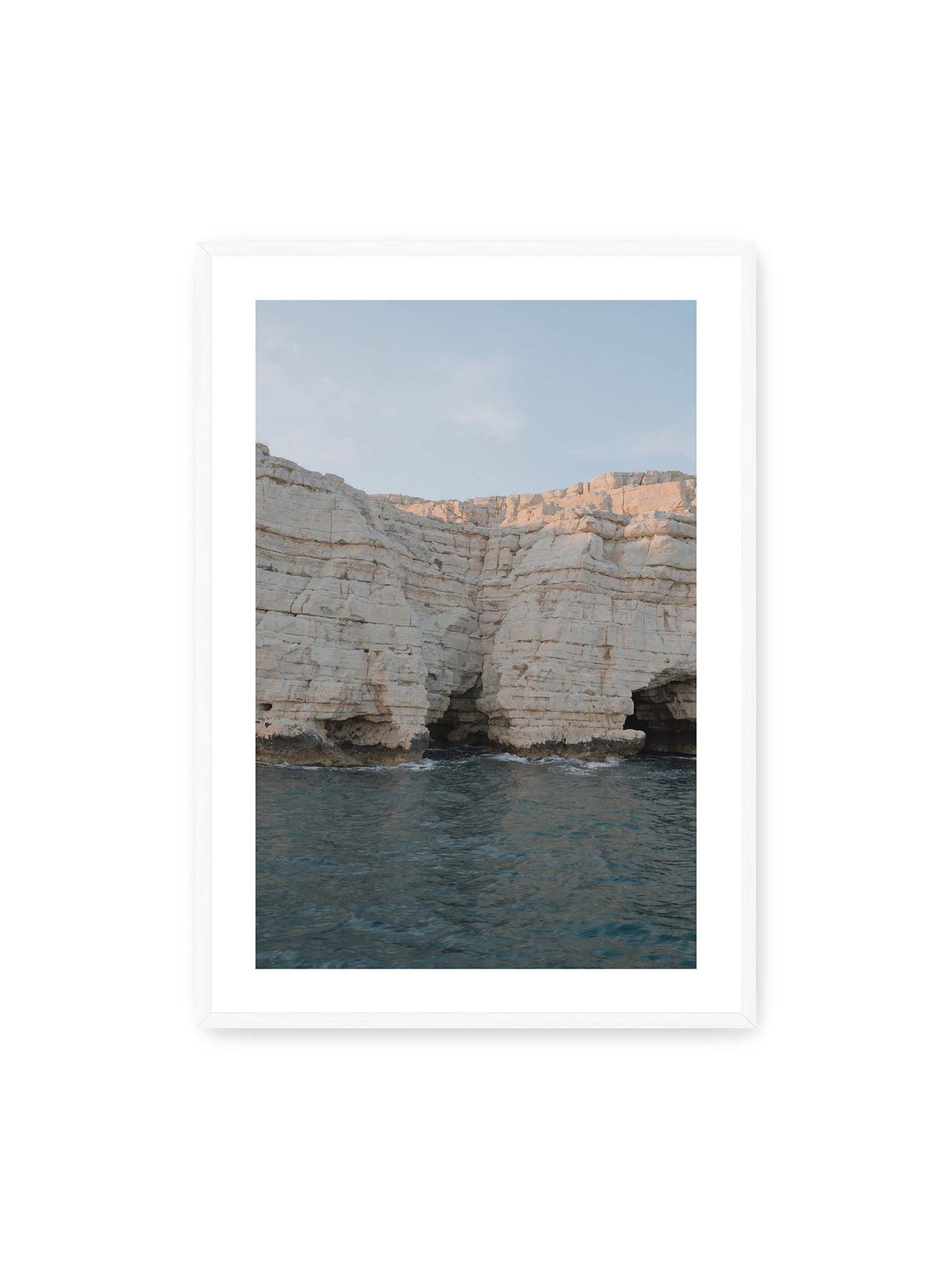 70x100 cm, weißer Galerie-Rand, weißer Rahmen – Helle geschichtete Felswand mit dunklen Grotten über bewegtem blauem Meer und blassem Himmel.