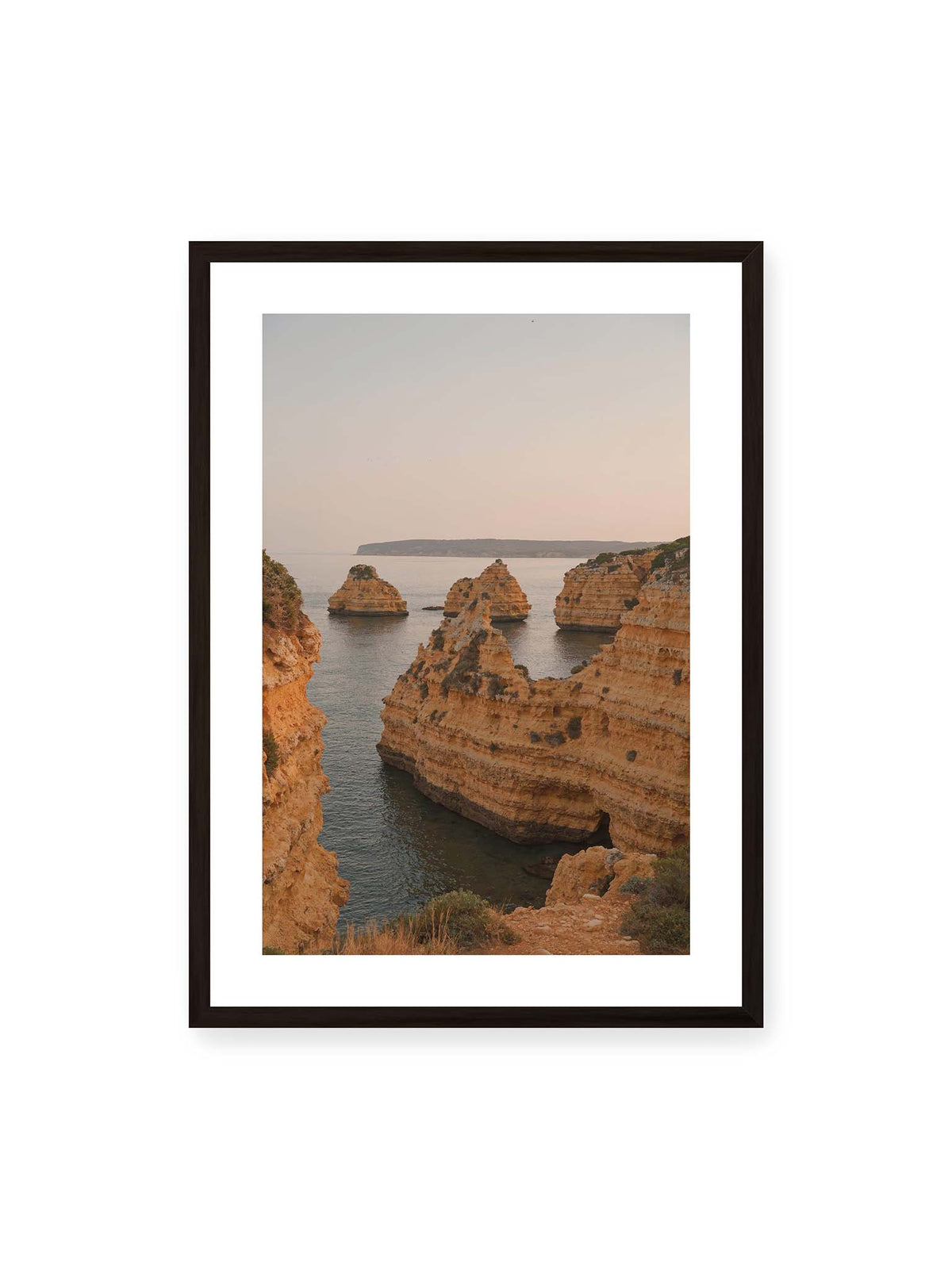 50x70 cm, weißer Galerie-Rand, schwarzer Rahmen – Warm getönte, geschichtete Felsformationen ragen zwischen steilen Küstenwänden aus ruhigem Meer.