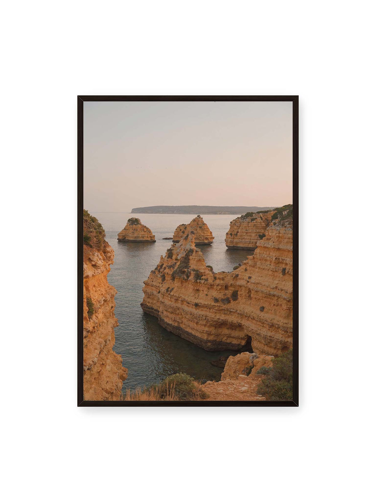 70x100 cm, schwarzer Rahmen – Warm getönte, geschichtete Felsformationen ragen zwischen steilen Küstenwänden aus ruhigem Meer.