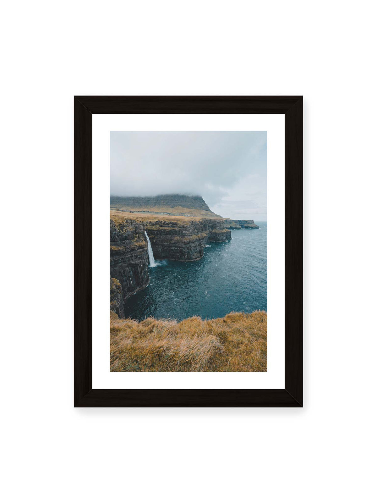 21x30 cm, weißer Galerie-Rand, schwarzer Rahmen – Dunkle Küstenklippen mit Wasserfall, grasigem Hochland und tief hängenden Wolken über blauem Meer.