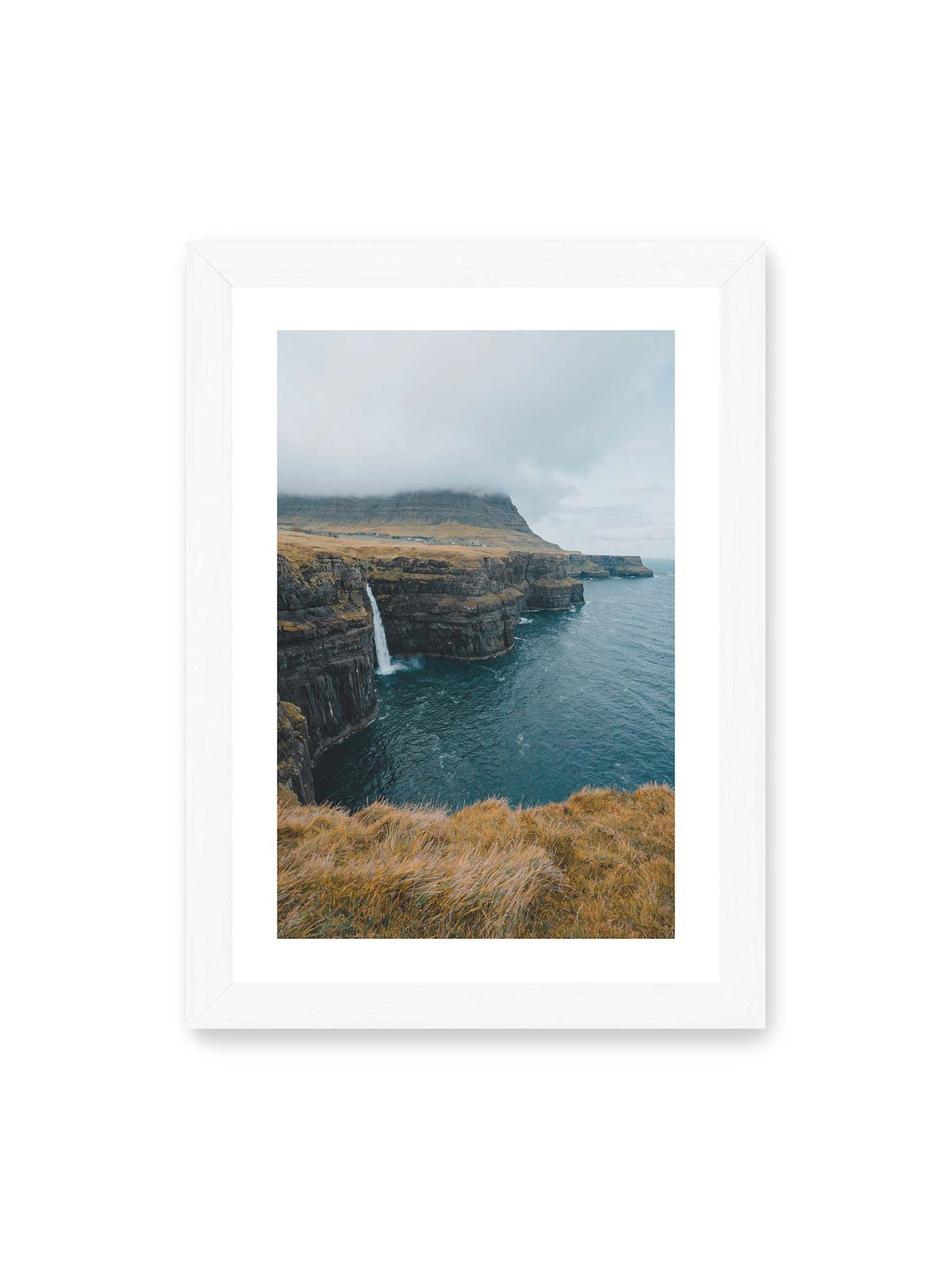 21x30 cm, weißer Galerie-Rand, weißer Rahmen – Dunkle Küstenklippen mit Wasserfall, grasigem Hochland und tief hängenden Wolken über blauem Meer.