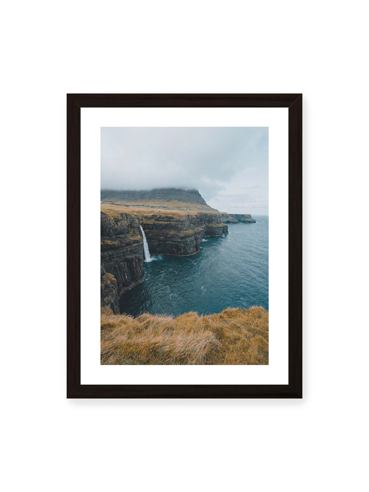 30x40 cm, weißer Galerie-Rand, schwarzer Rahmen – Dunkle Küstenklippen mit Wasserfall, grasigem Hochland und tief hängenden Wolken über blauem Meer.