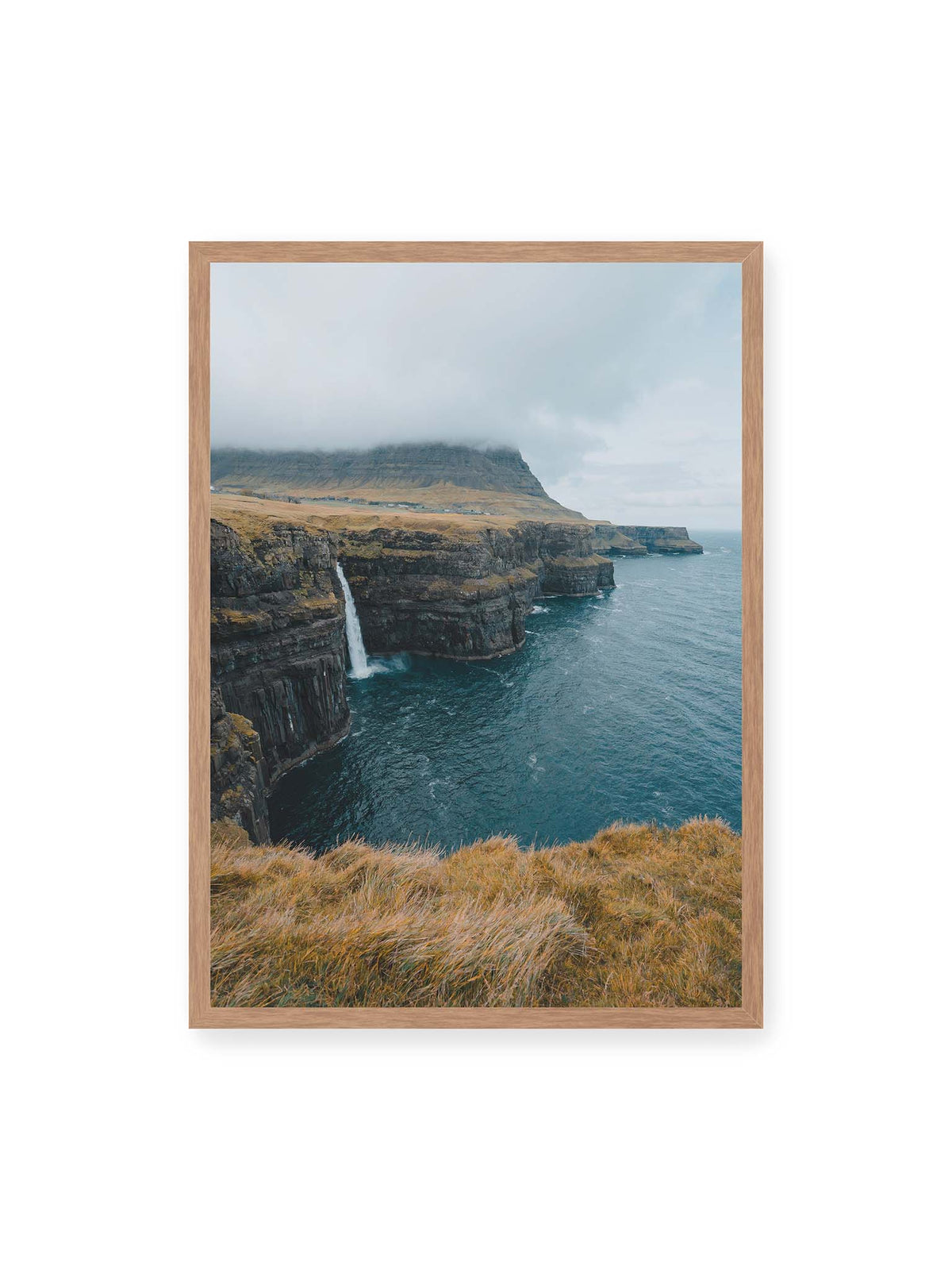50x70 cm, Eichenrahmen – Dunkle Küstenklippen mit Wasserfall, grasigem Hochland und tief hängenden Wolken über blauem Meer.