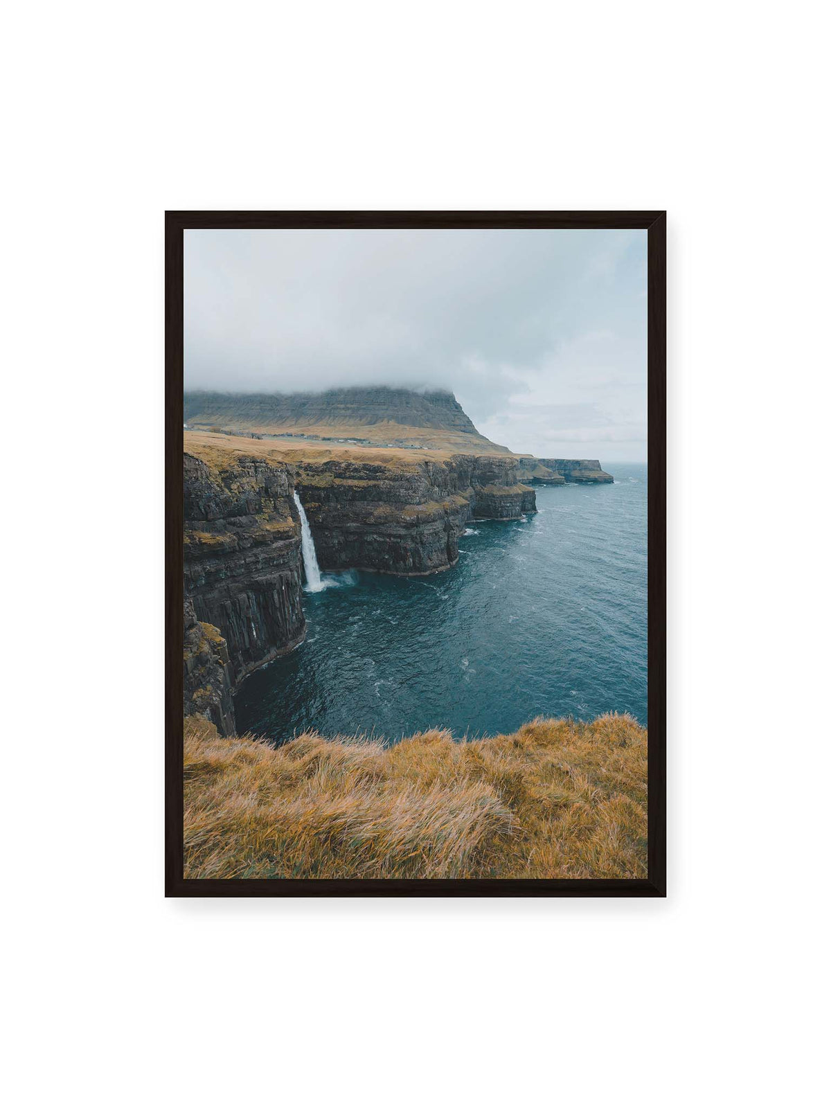 50x70 cm, schwarzer Rahmen – Dunkle Küstenklippen mit Wasserfall, grasigem Hochland und tief hängenden Wolken über blauem Meer.