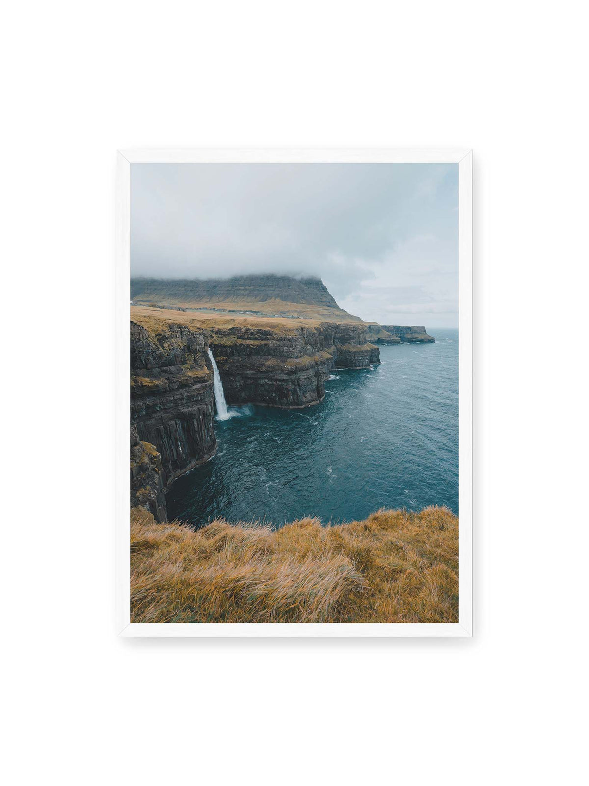 50x70 cm, weißer Rahmen – Dunkle Küstenklippen mit Wasserfall, grasigem Hochland und tief hängenden Wolken über blauem Meer.